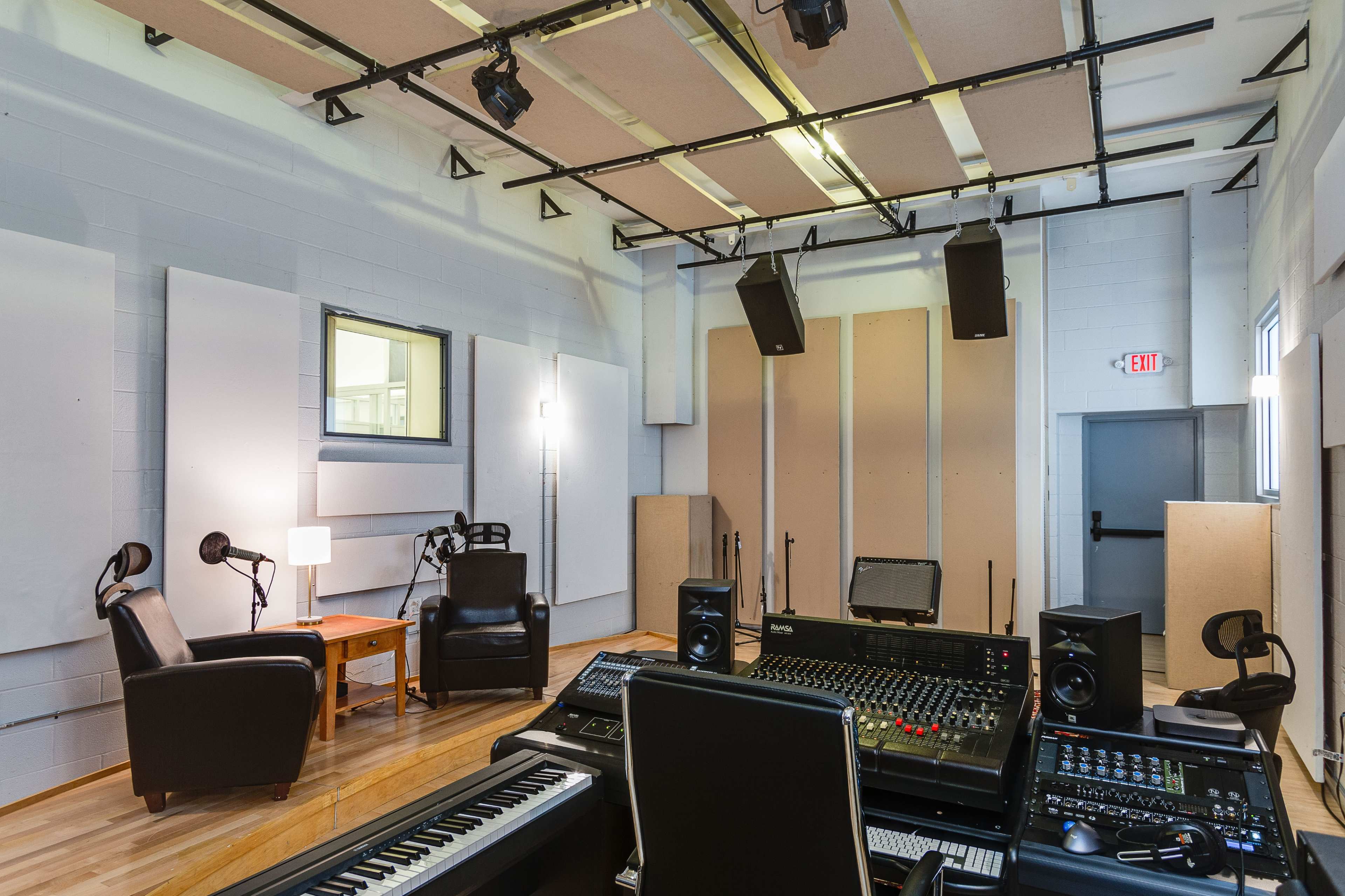 A sound recording studio featuring a control console, acoustic panels on the walls, two chairs, a side table, and overhead speakers.