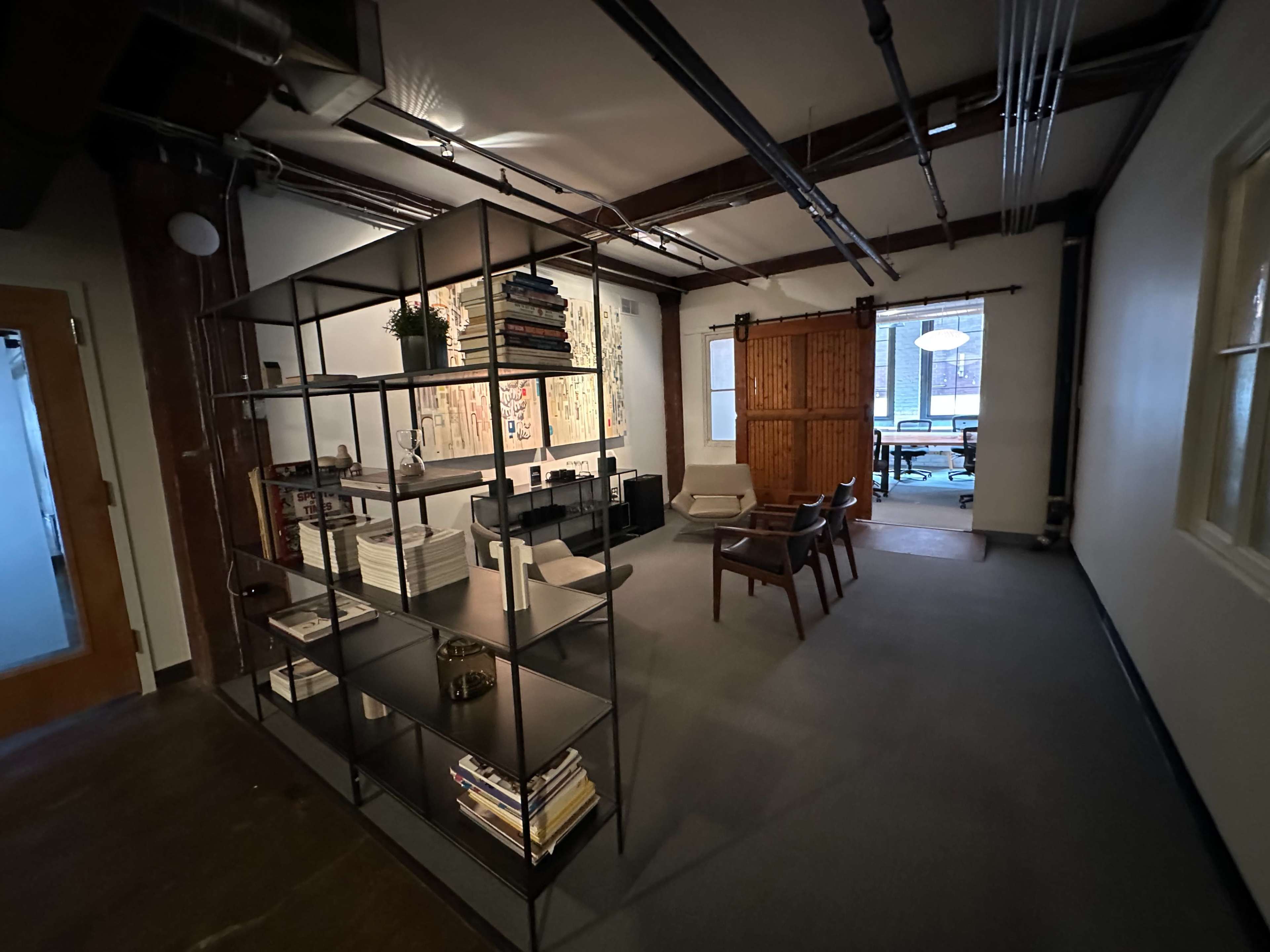 A modern office space featuring a metal bookshelf divider, a seating area with chairs, and large windows letting in natural light.