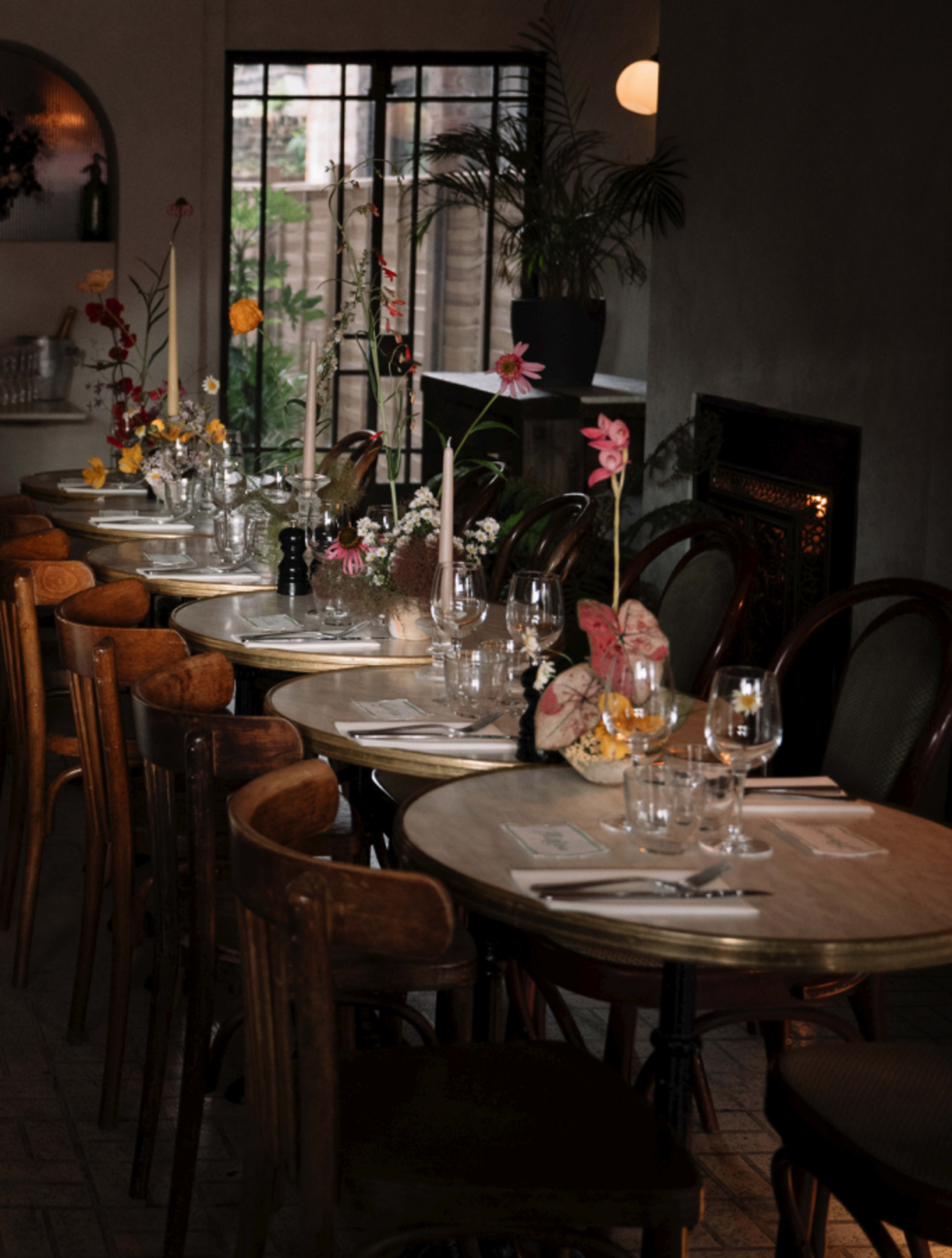 A row of elegantly set tables with glassware and floral centerpieces is arranged in a dimly lit restaurant.