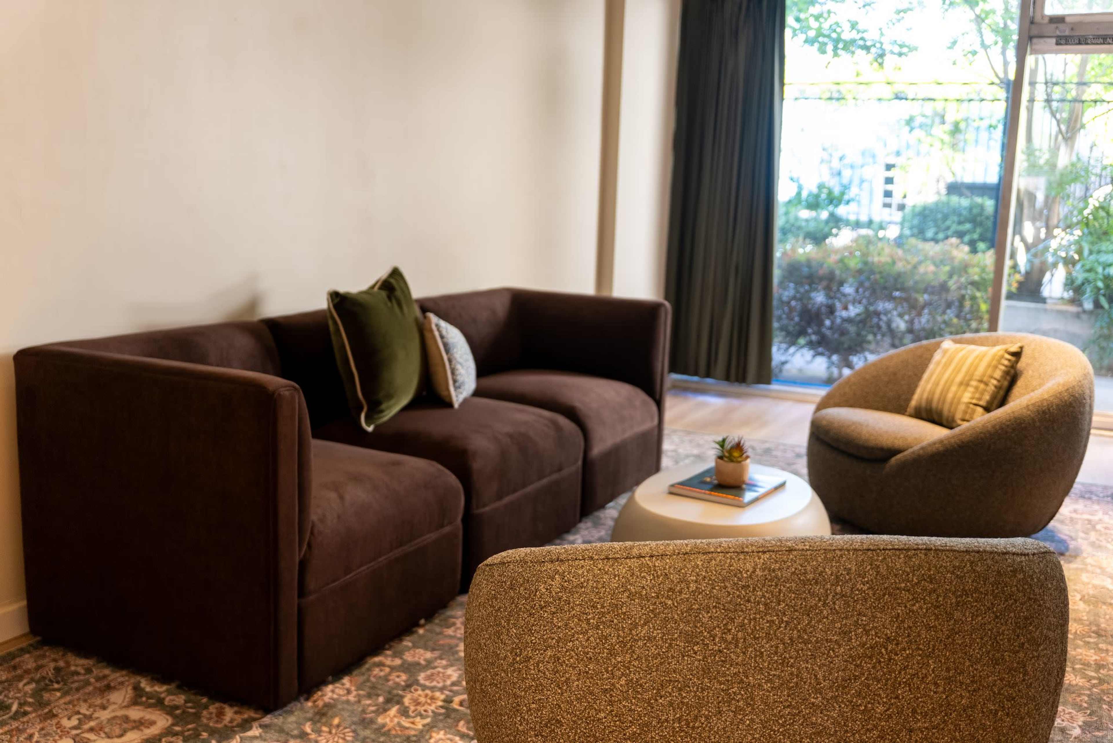 The image shows a modern living space featuring a brown upholstered sofa, a round coffee table with a small plant, and a gray accent chair near a large window.