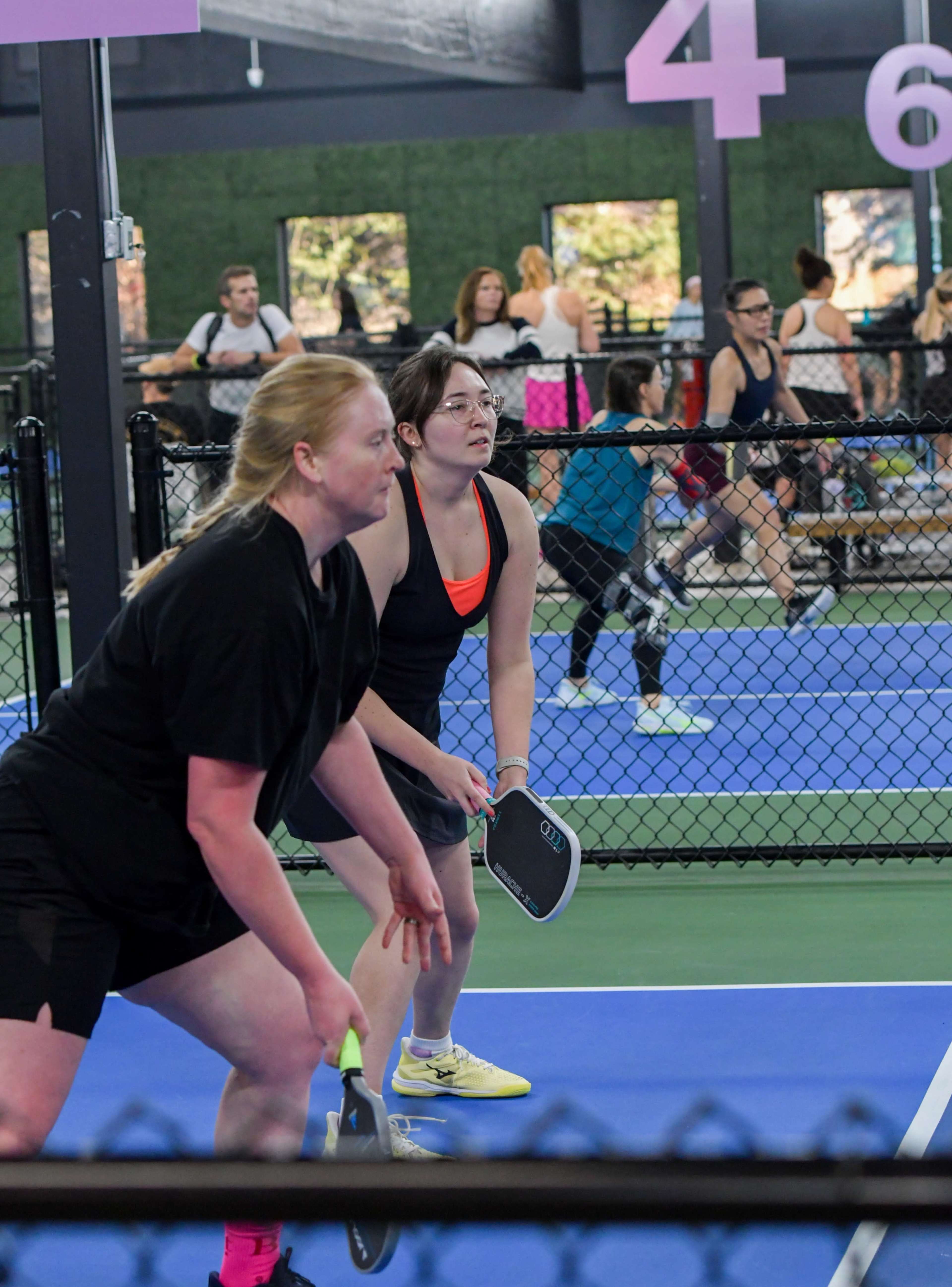 Two women prepare to serve in a pickleball match while others play in the background at an indoor sports facility.
