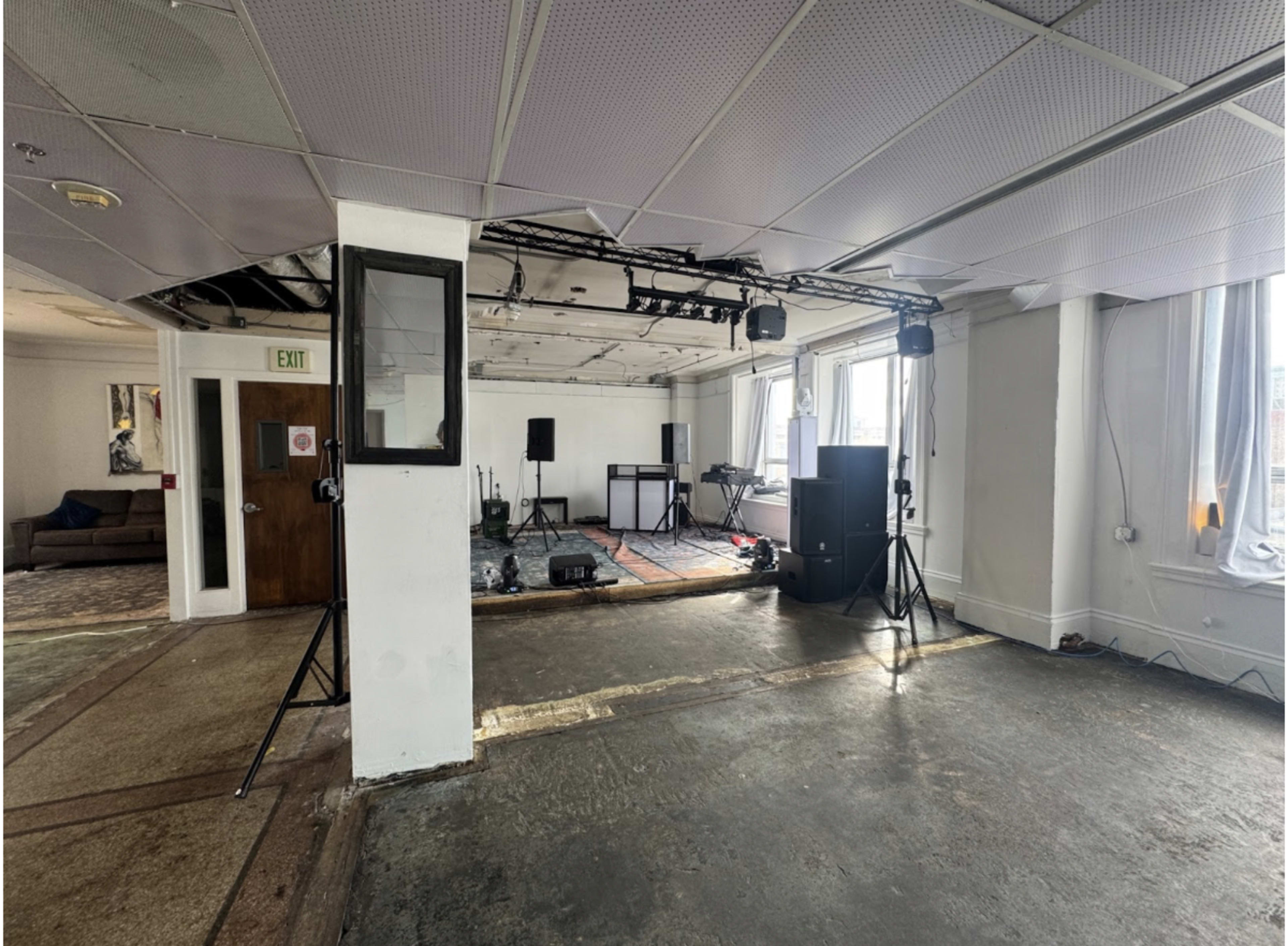 The image shows a large, empty room with a bare floor, stage equipment on one end, and several light fixtures installed overhead.