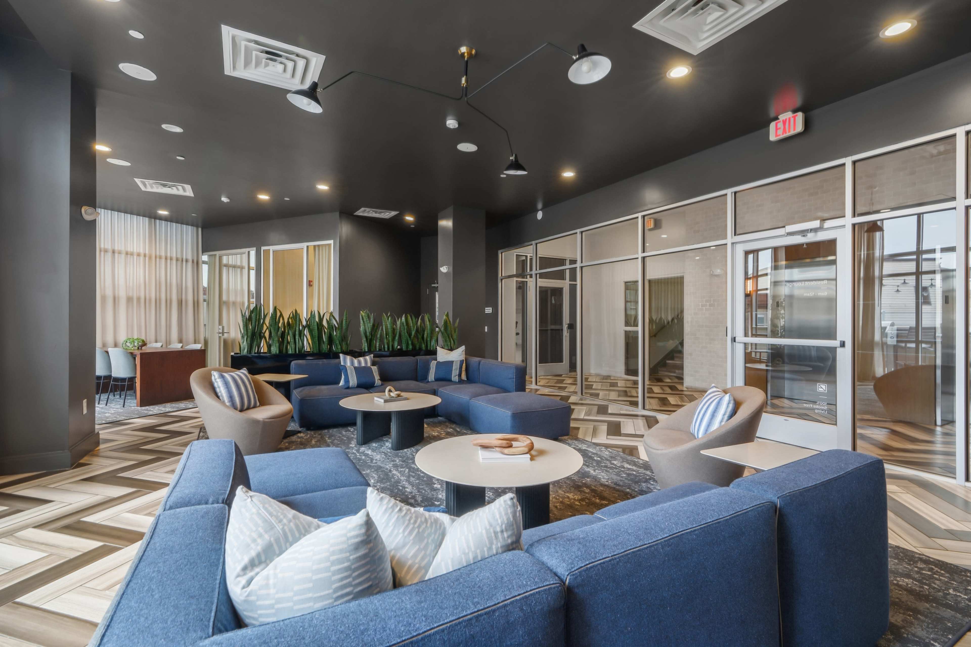 The image shows a modern lounge area featuring blue sofas, round tables, and greenery, with large windows and contemporary lighting.