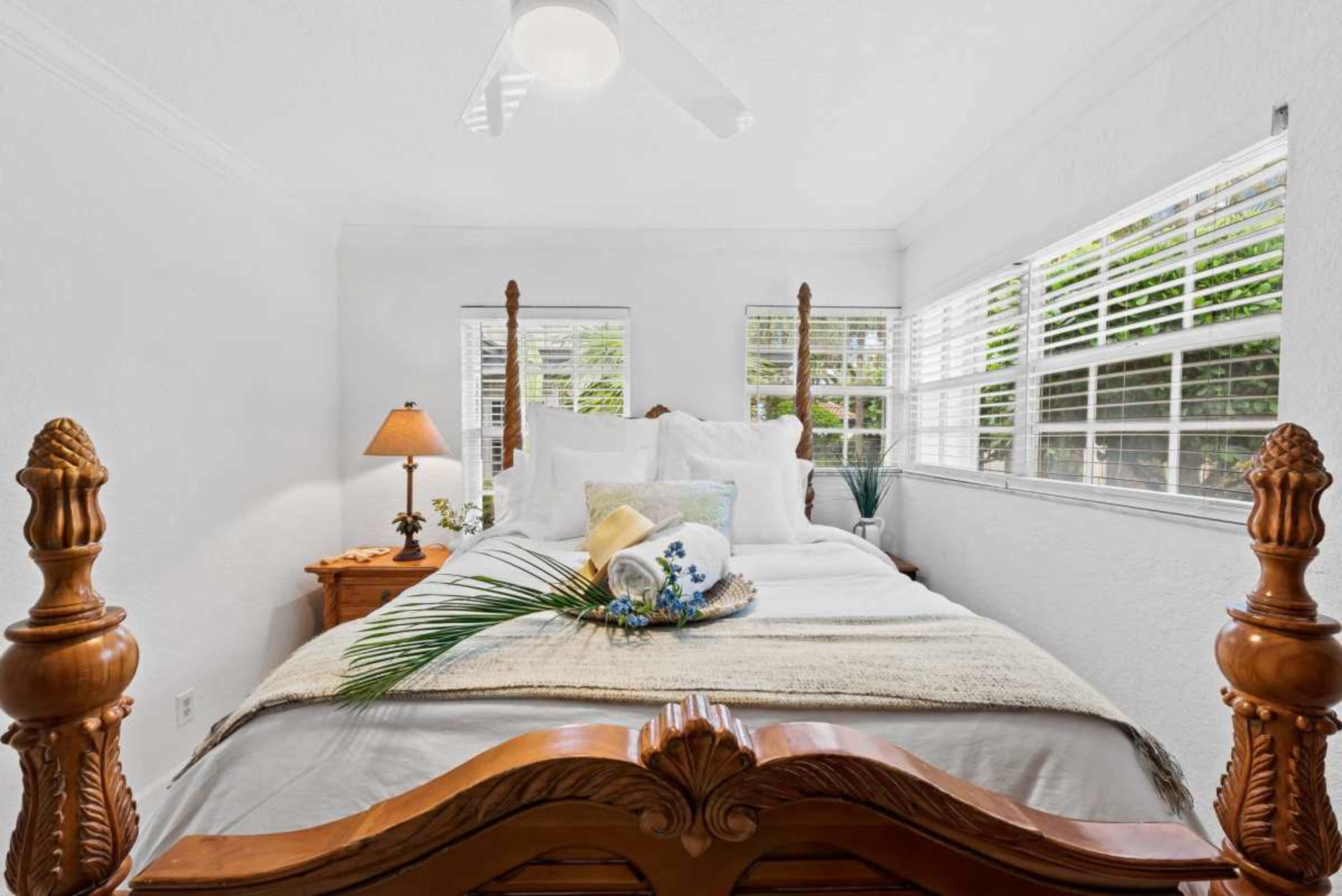 A neatly arranged bedroom features a wooden bed frame, white bedding, and sunlight streaming through window blinds.