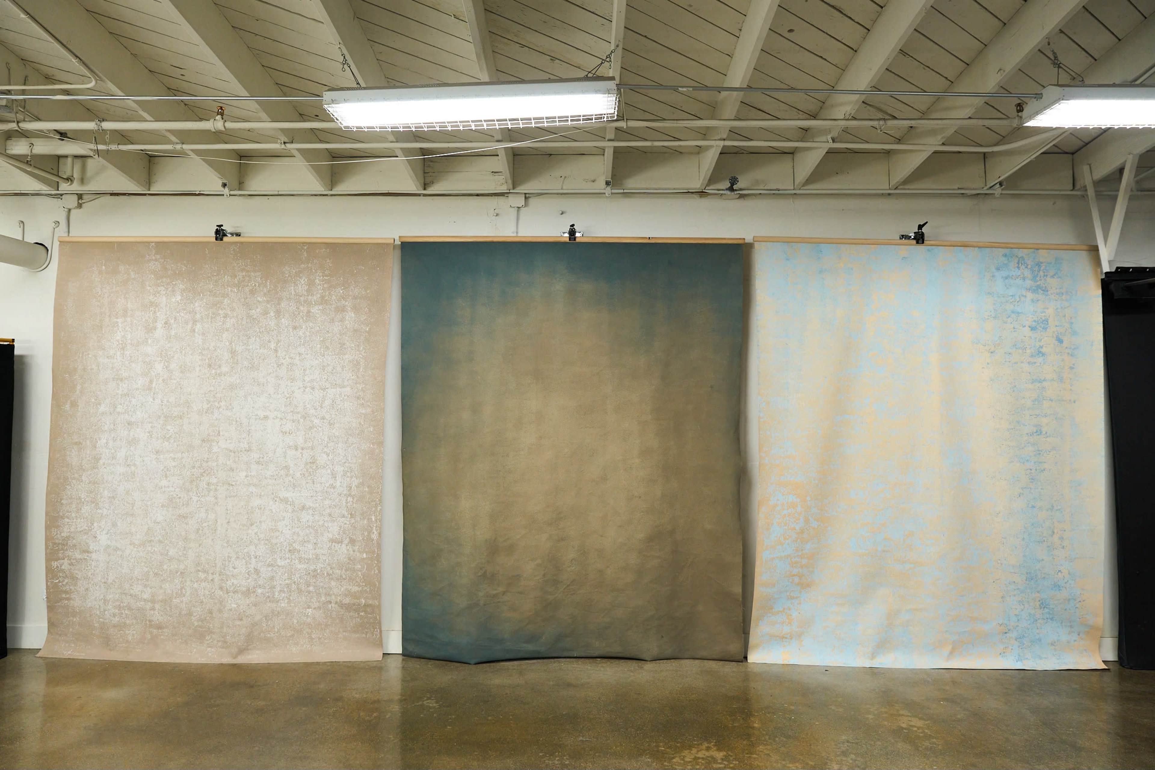 Three large fabric panels hanging against a wall in a room with a high ceiling, featuring varying shades of beige, brown, and pale blue.