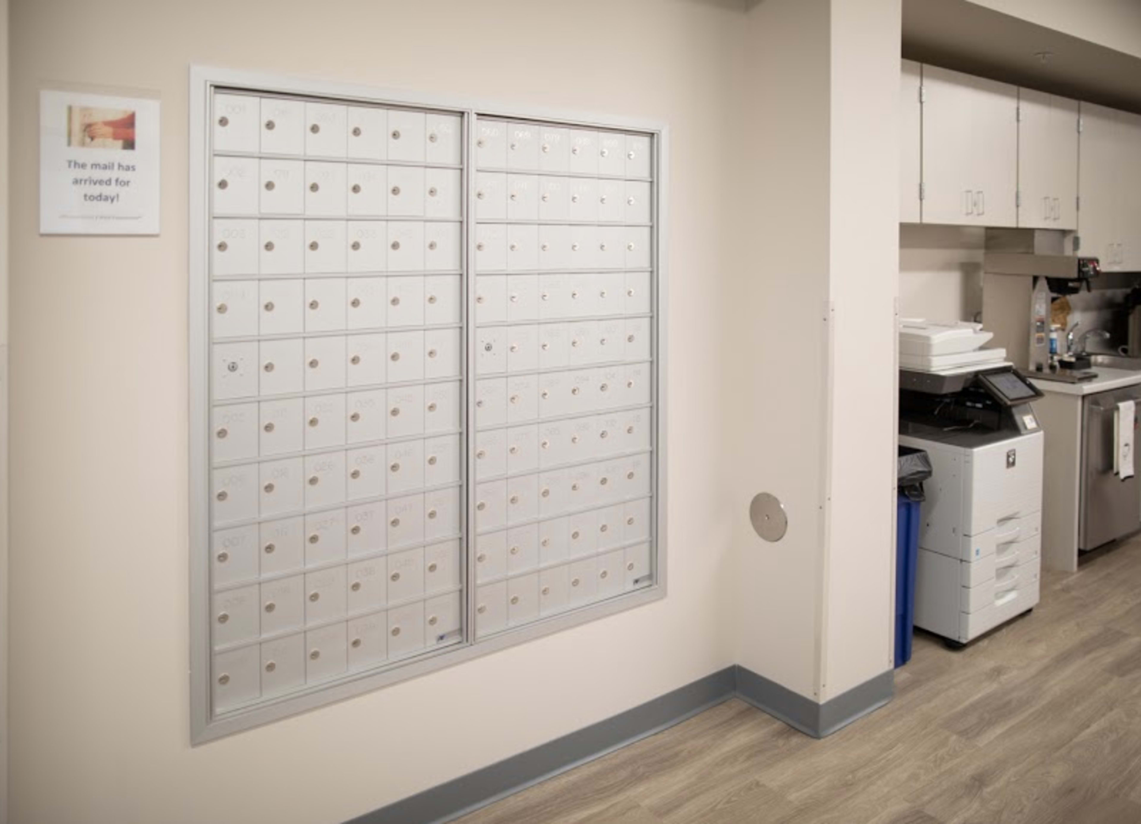 A mailroom features a wall with a grid of small mailboxes and a sign indicating that mail has arrived, alongside a printer and trash receptacle.