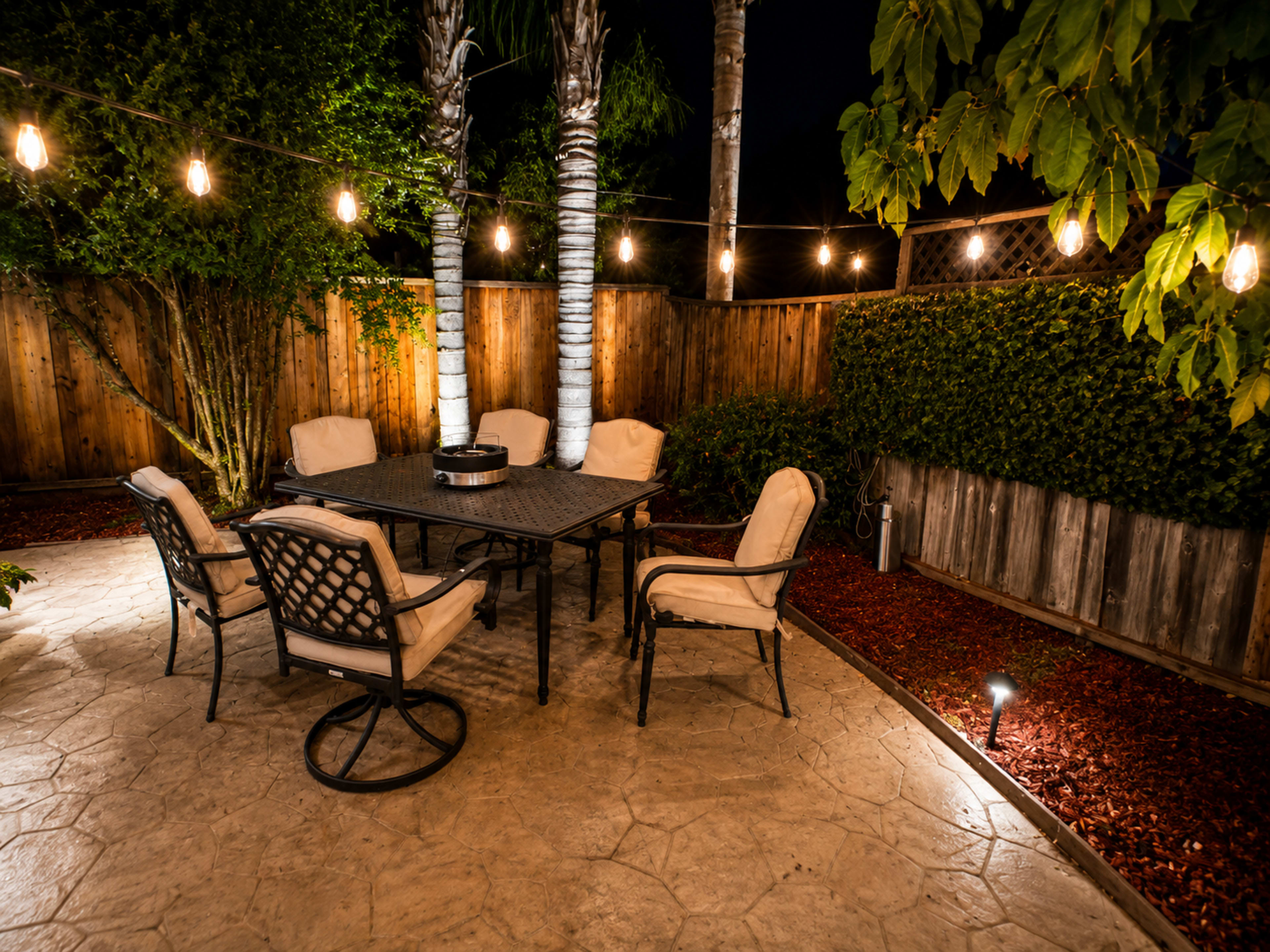 The image shows an outdoor dining area at night, featuring a round table surrounded by six chairs, with string lights illuminating the space and palm trees in the background.