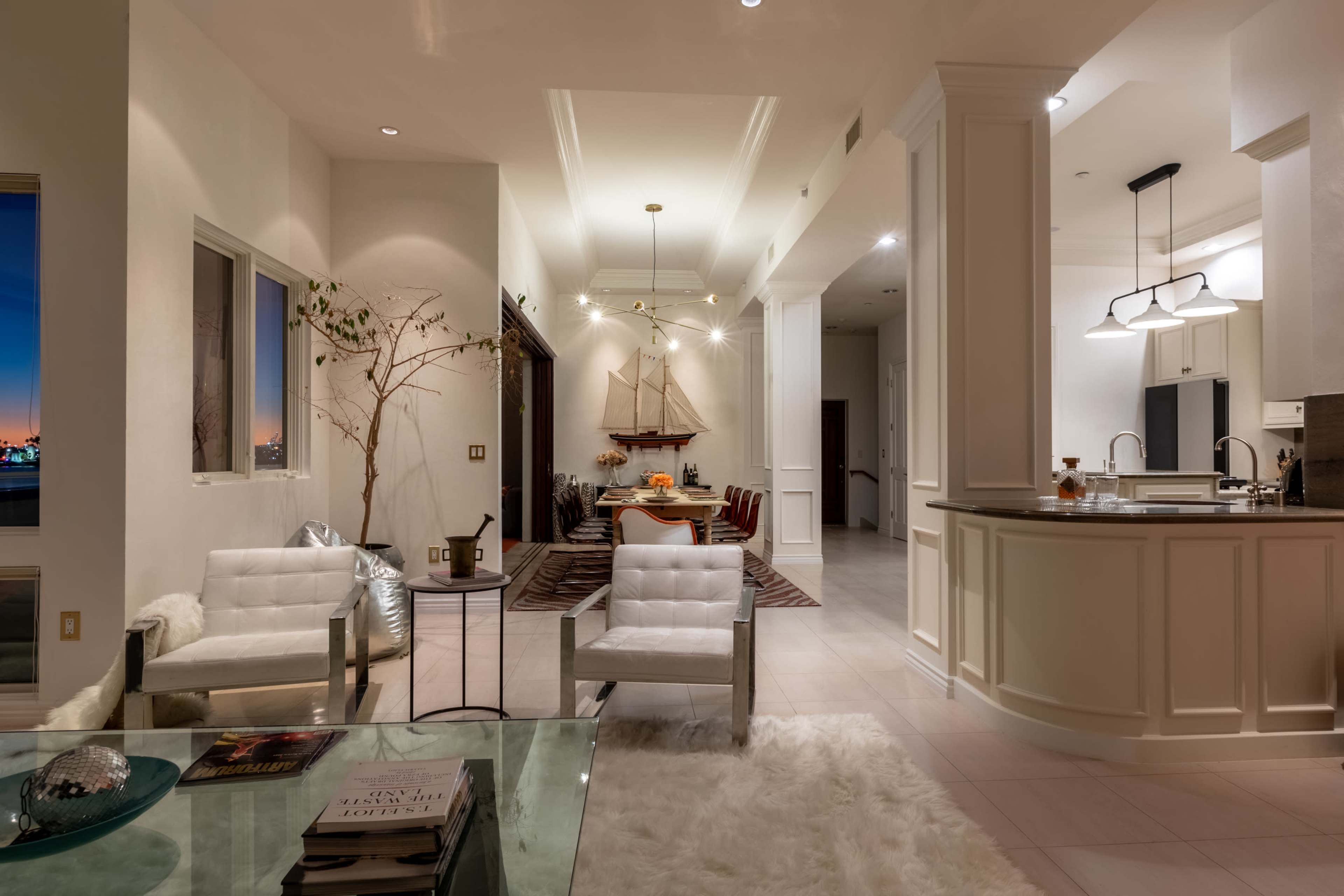 The image shows a modern, open-concept living space with a kitchen, dining area, and seating area featuring contemporary furniture and decorative elements.