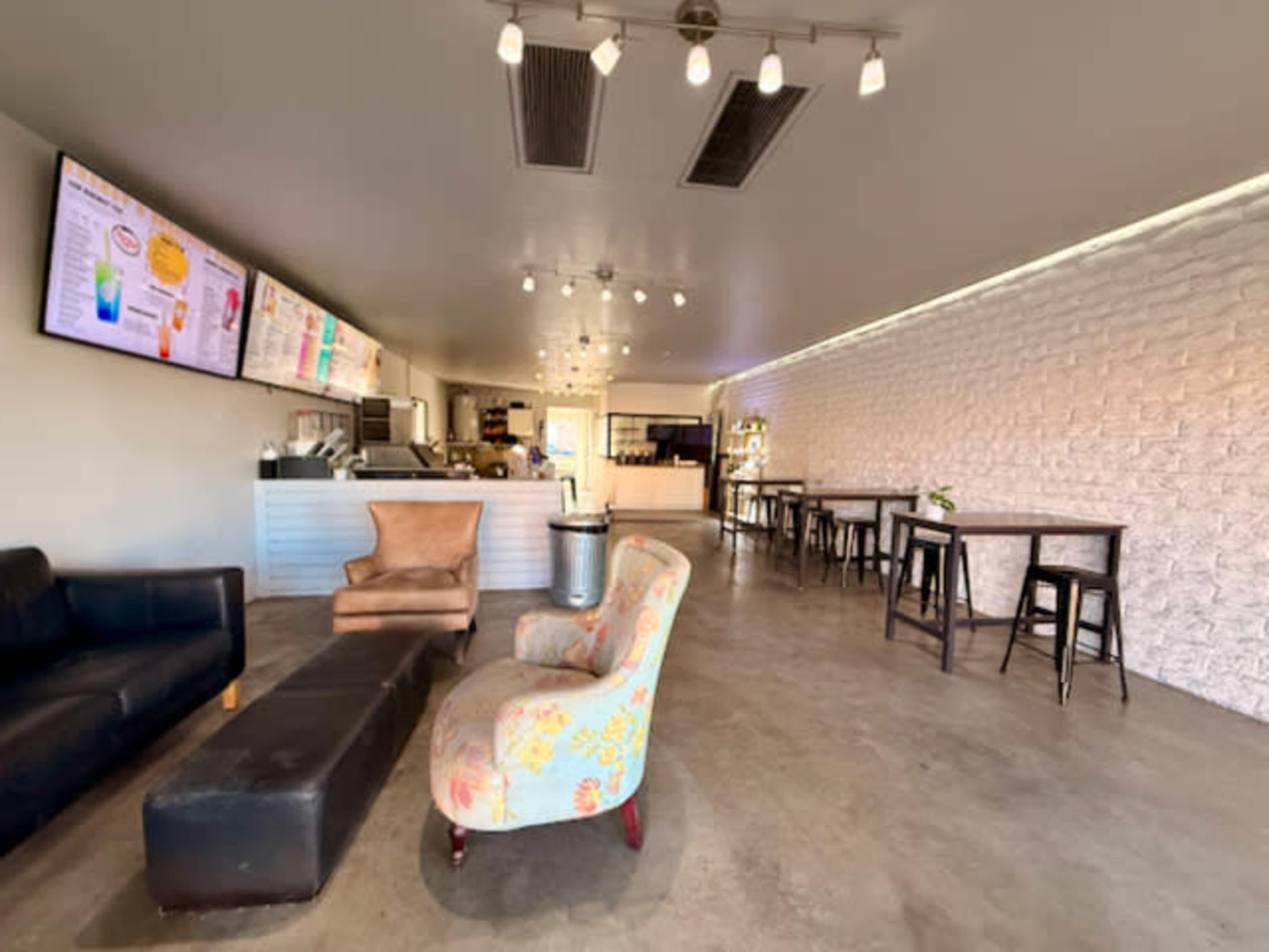 The image shows a modern coffee shop interior featuring a seating area with colorful chairs, a long black bench, and digital menu displays on the wall.