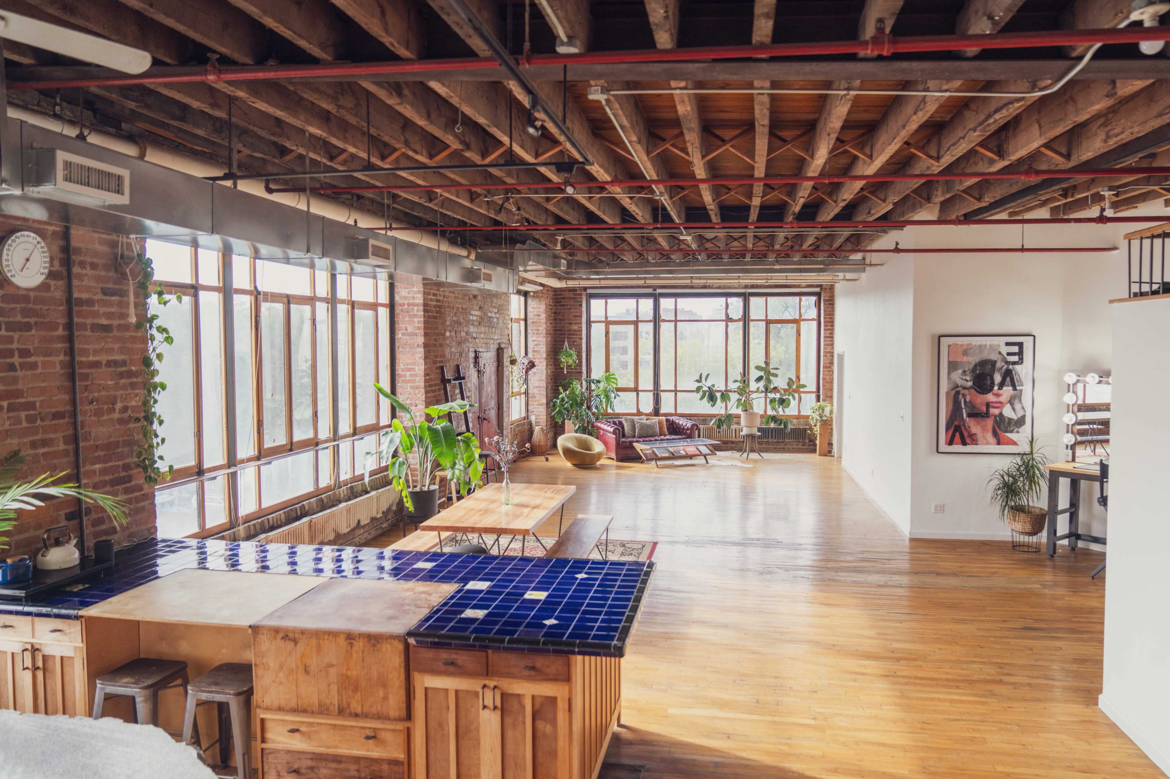 The image shows a spacious, loft-style living area with exposed wooden beams, large windows, and various plants scattered throughout.