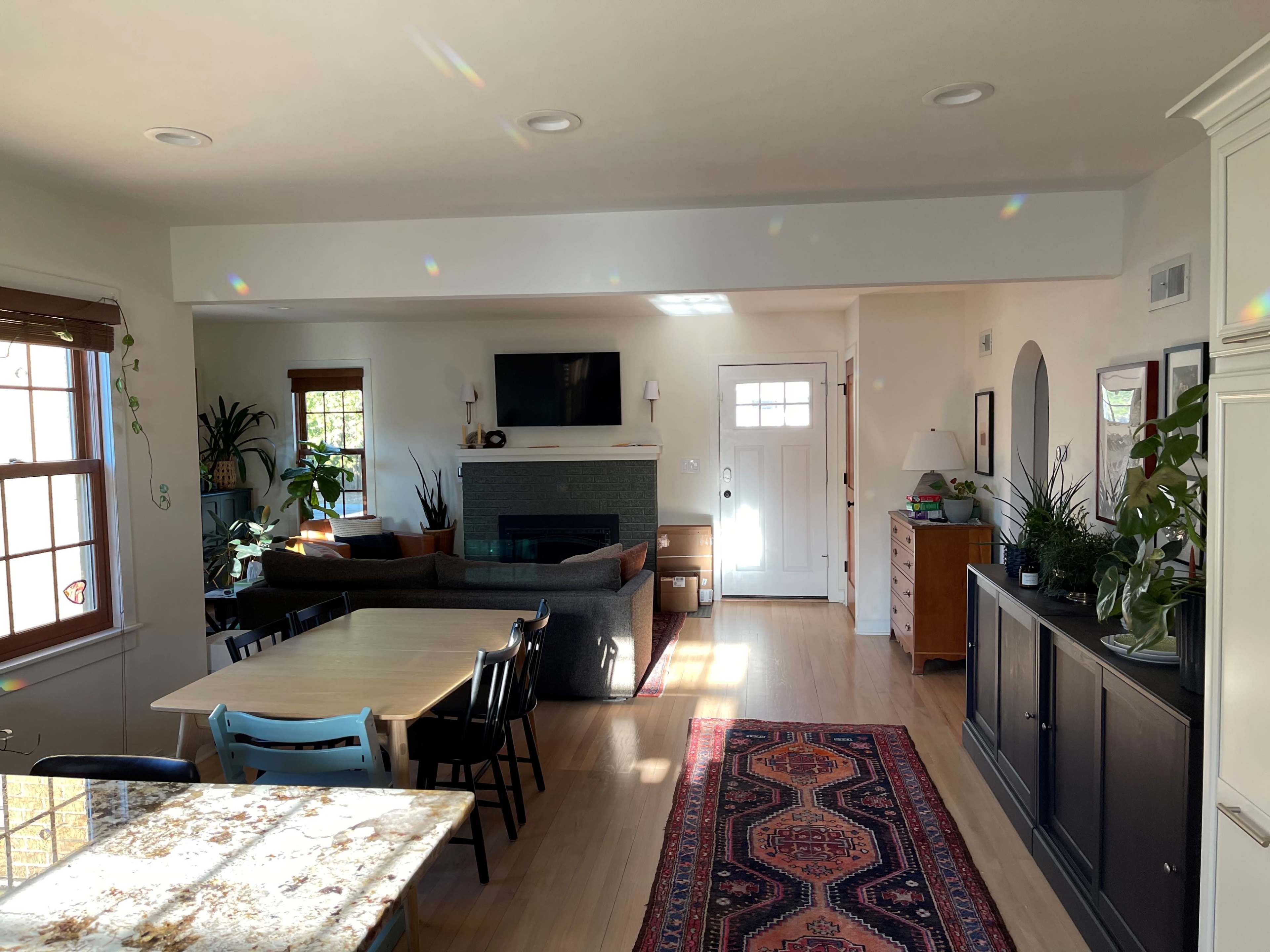 A spacious living area features a fireplace, a large window, a dining table, and various plants, with a front door visible in the background.