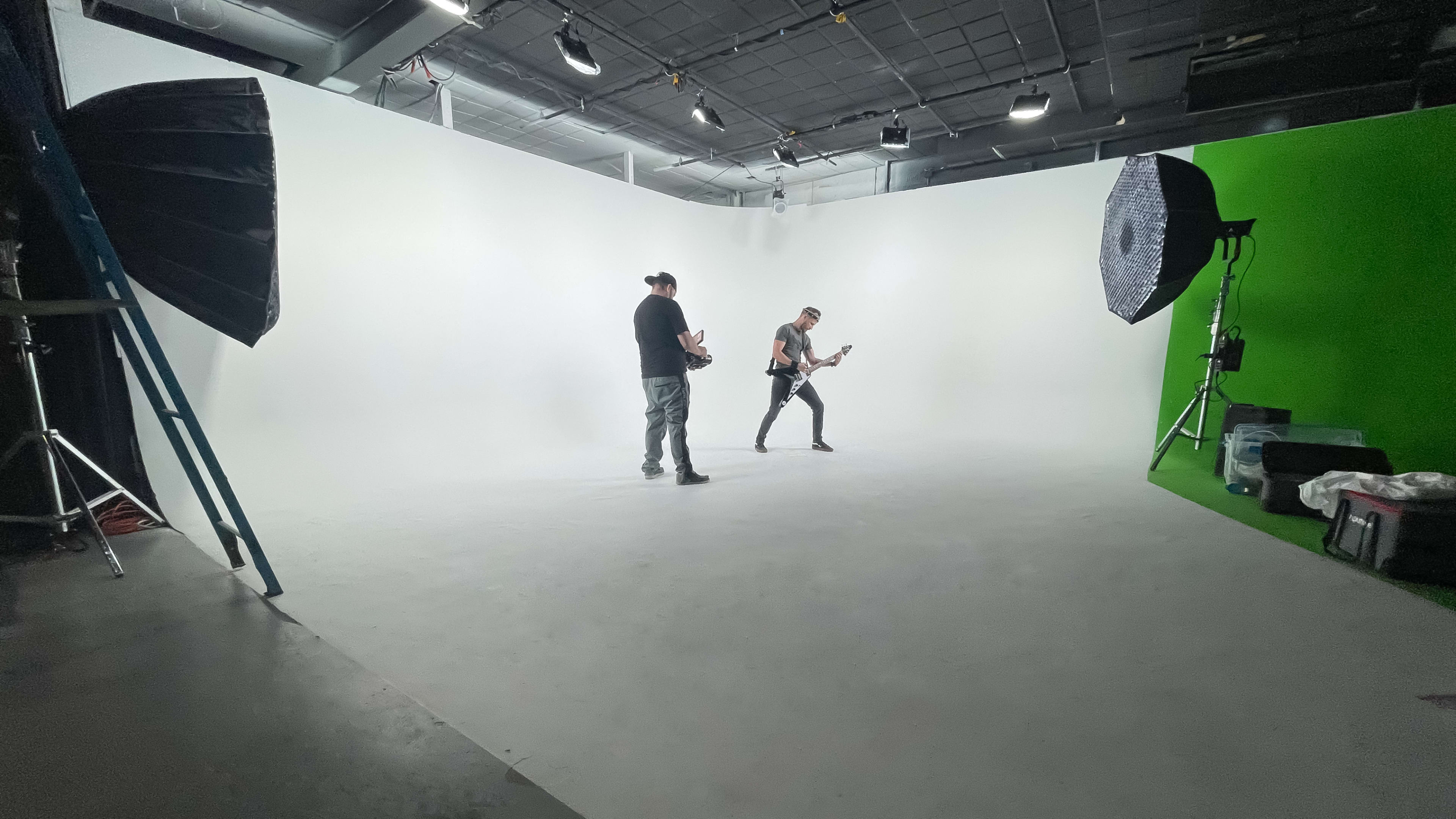 A musician is playing guitar on a white backdrop while another person stands nearby in a performance studio.