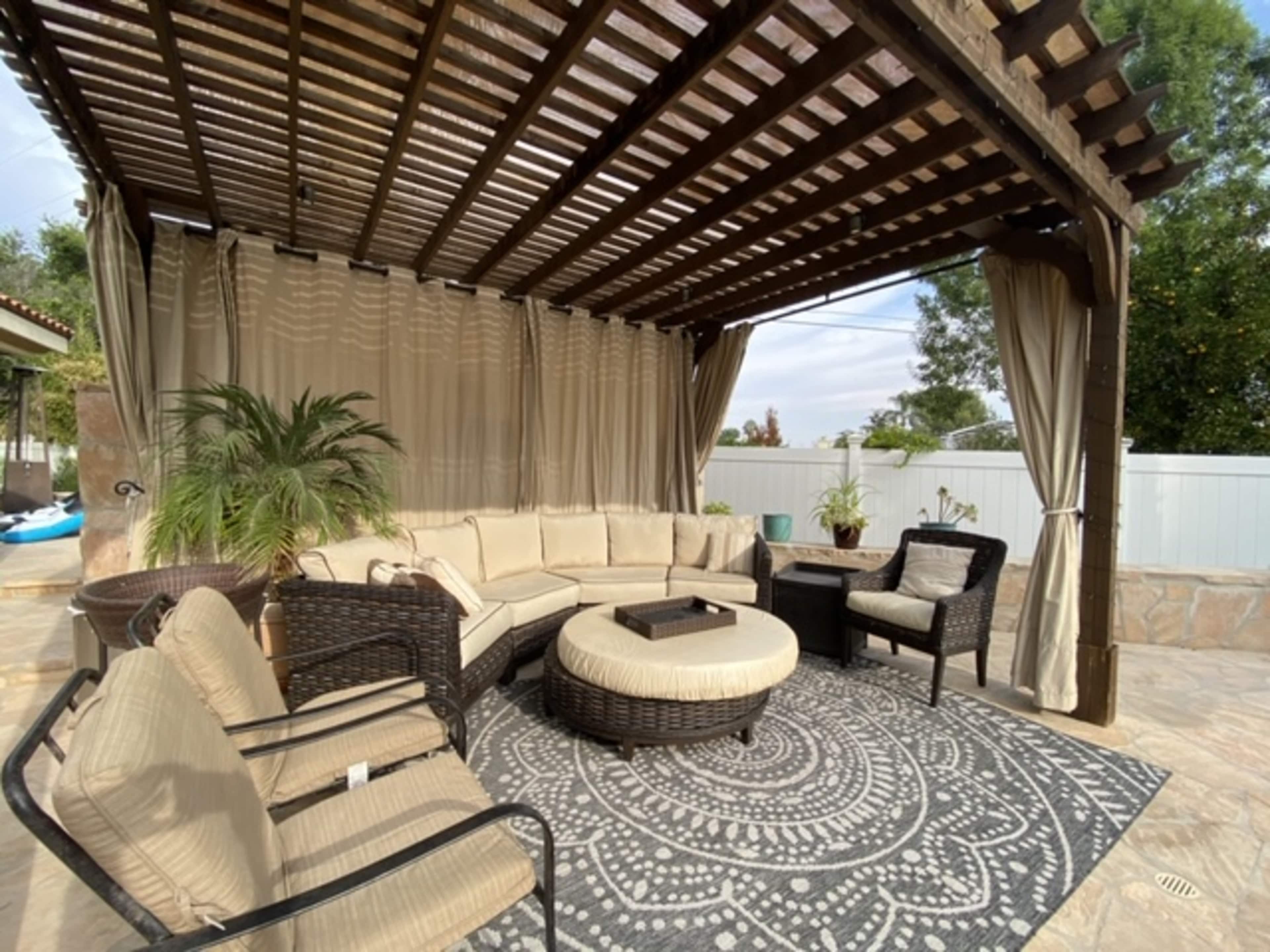 A covered outdoor seating area features cushioned furniture arranged around a central ottoman on a patterned rug.