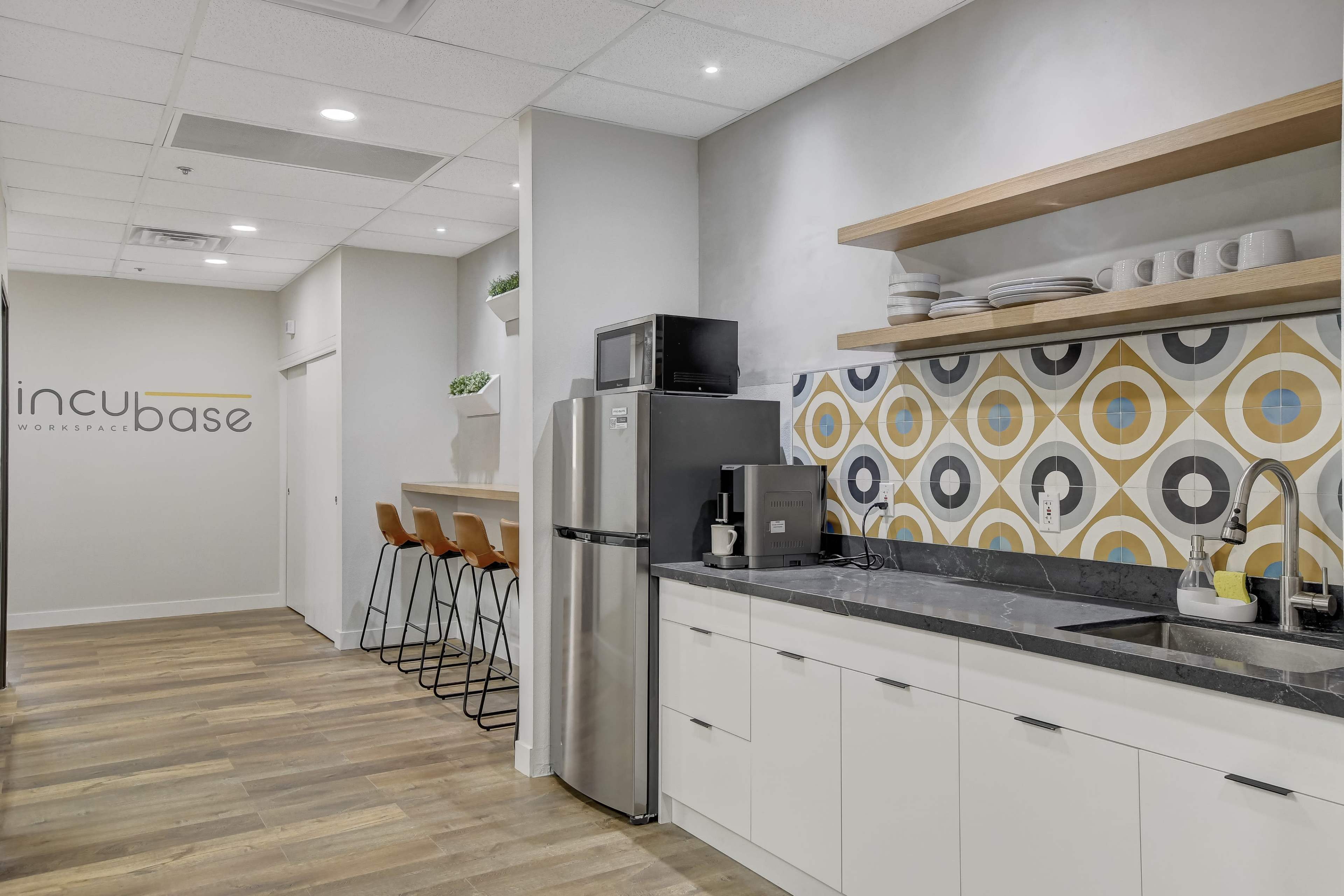 A modern kitchen area in a workspace, featuring a stainless steel fridge, a coffee machine, and patterned wall tiles, with bar stools positioned at a countertop.