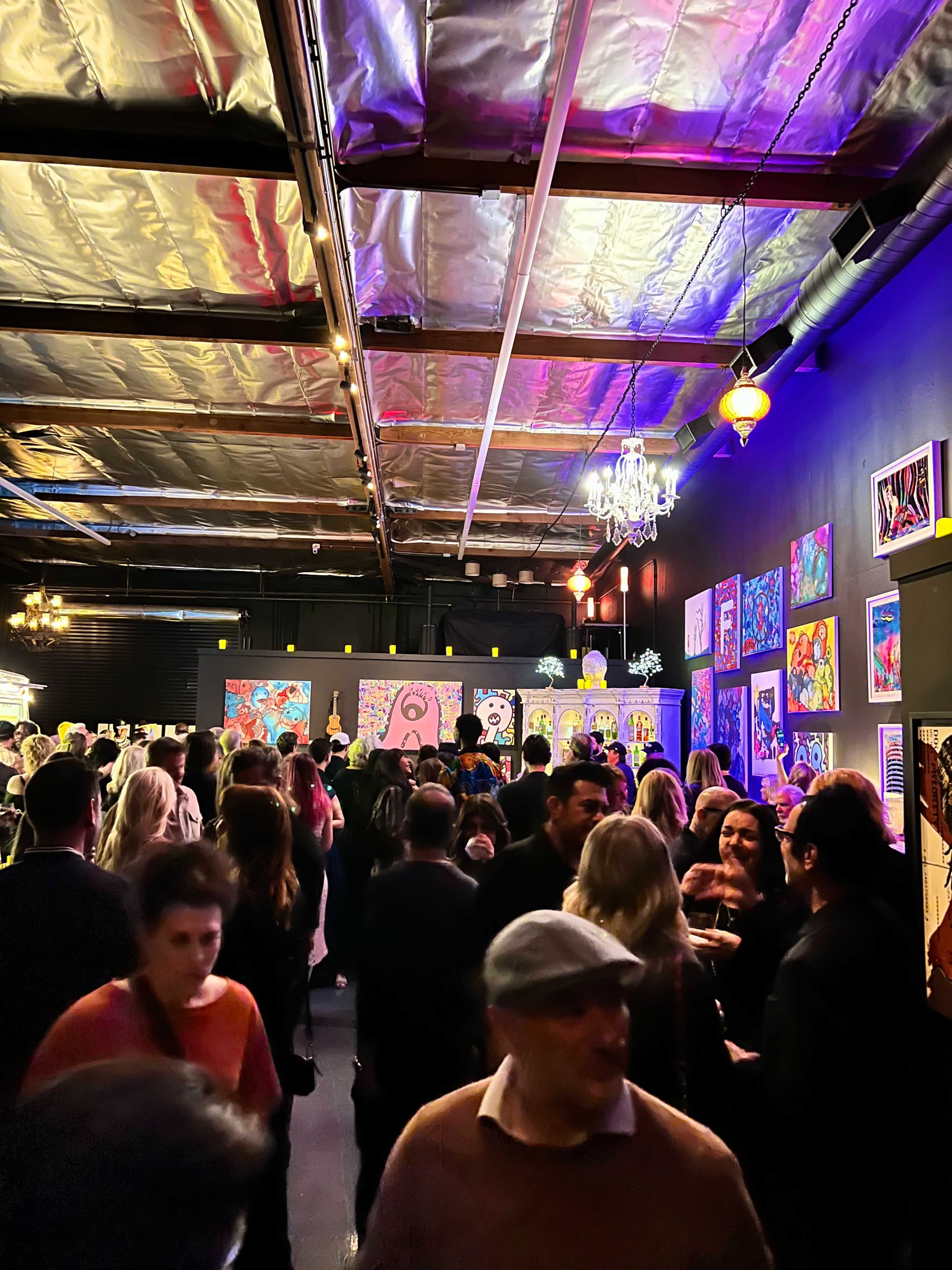 A crowd of people mingles in a brightly lit art gallery filled with colorful paintings on the walls.