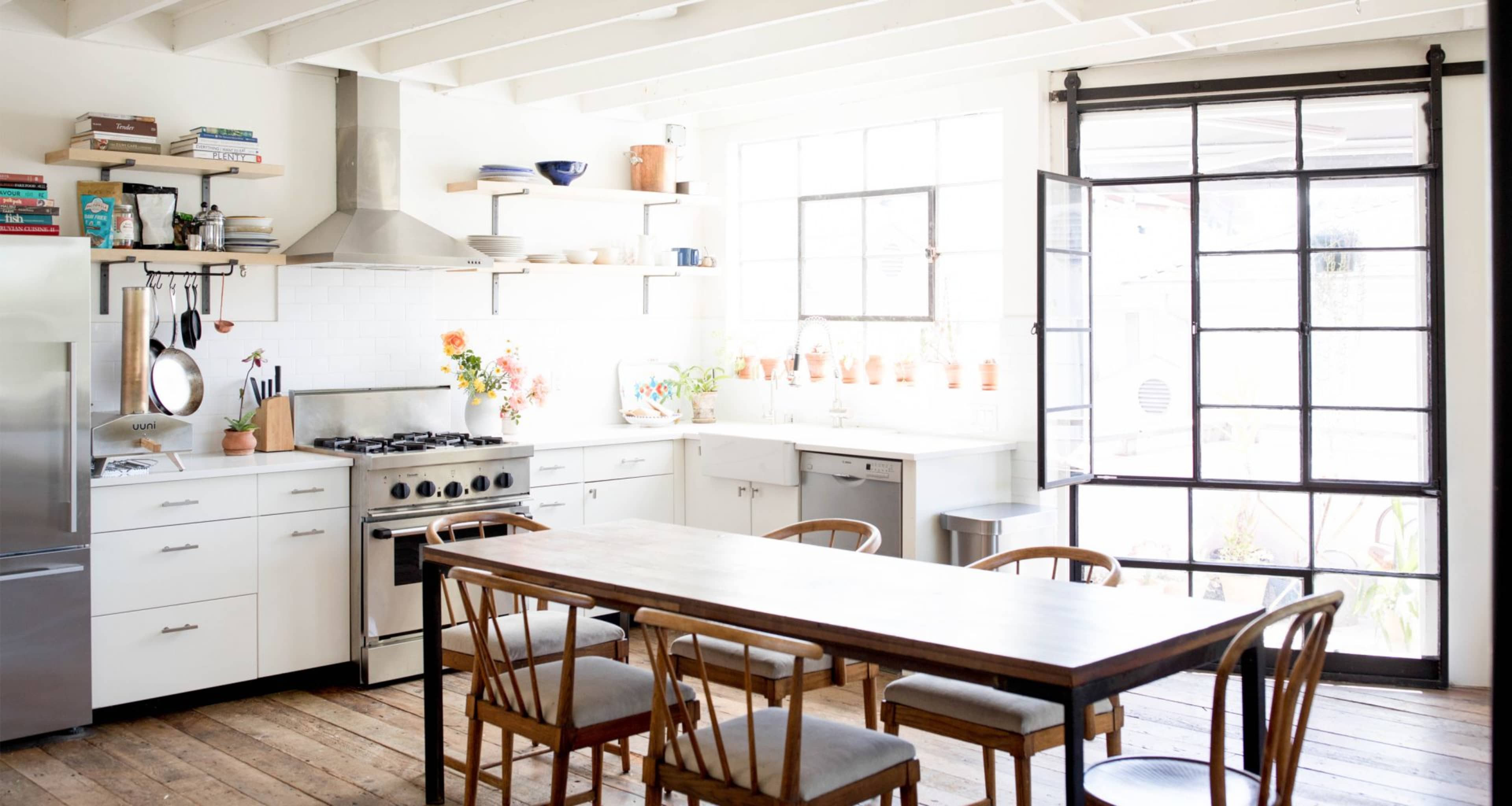 A bright, open kitchen featuring a central wooden dining table, modern appliances, and large windows with plants and decorative items on the shelves.