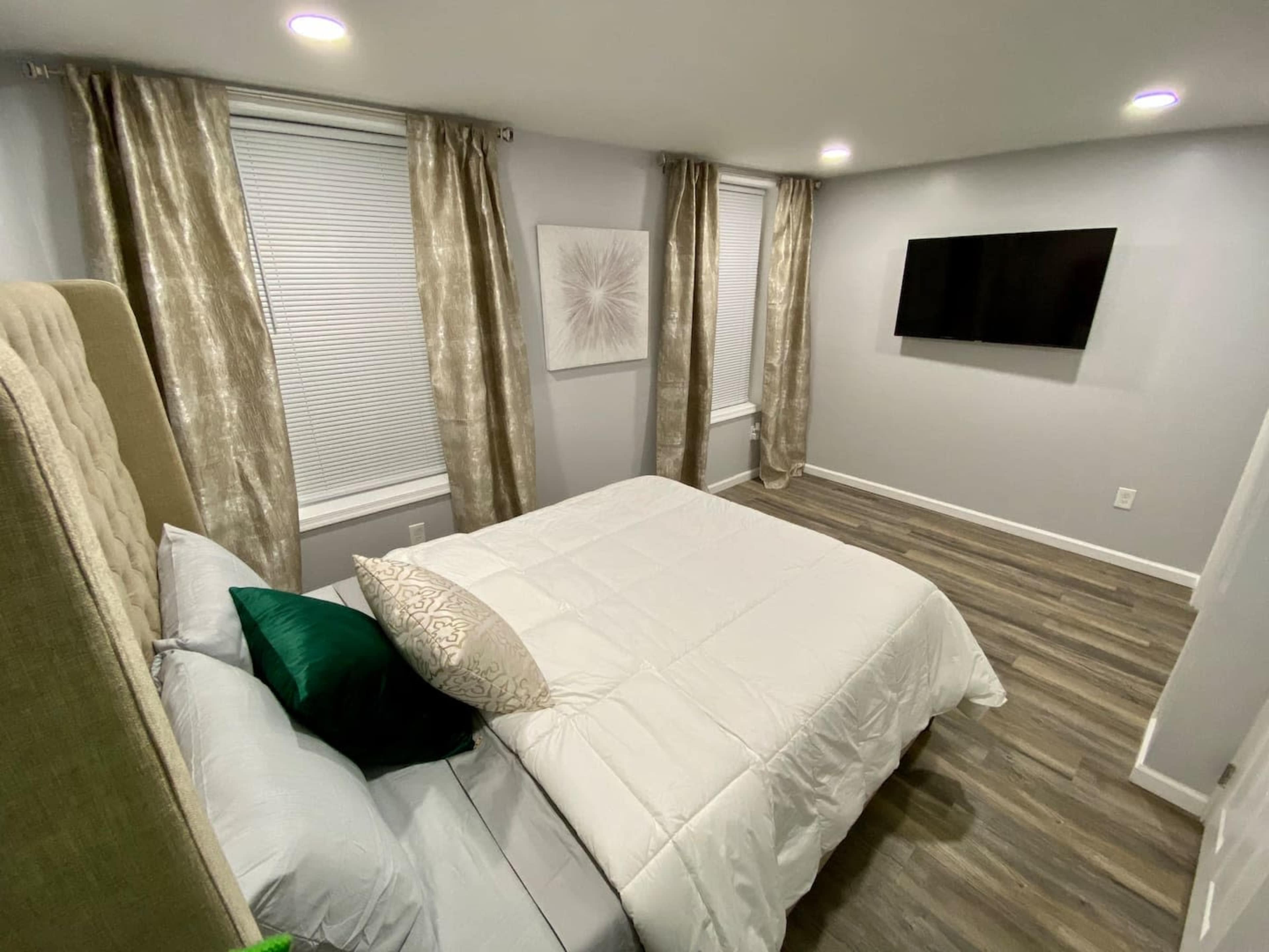 A modern bedroom with a bed, decorative pillows, a wall-mounted TV, and curtains covering two windows.