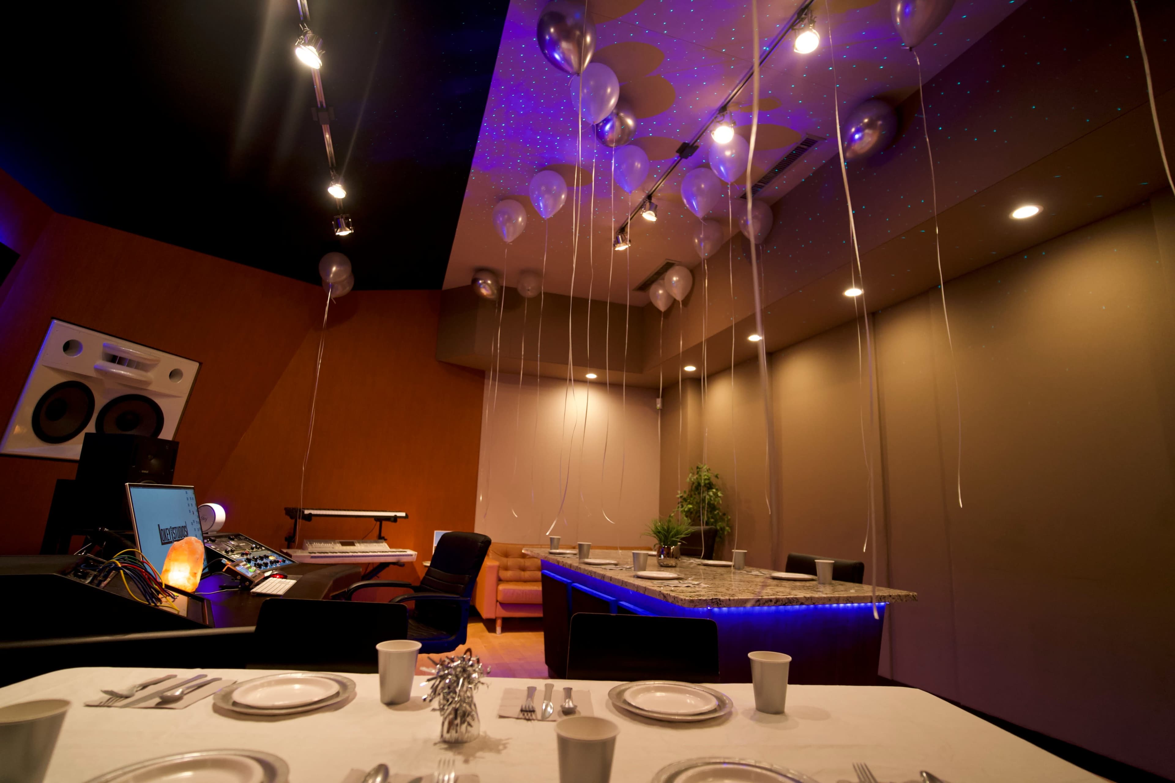 A recording studio is set up for a celebration, featuring a dining table with plates and cups, surrounded by balloons suspended from the ceiling.