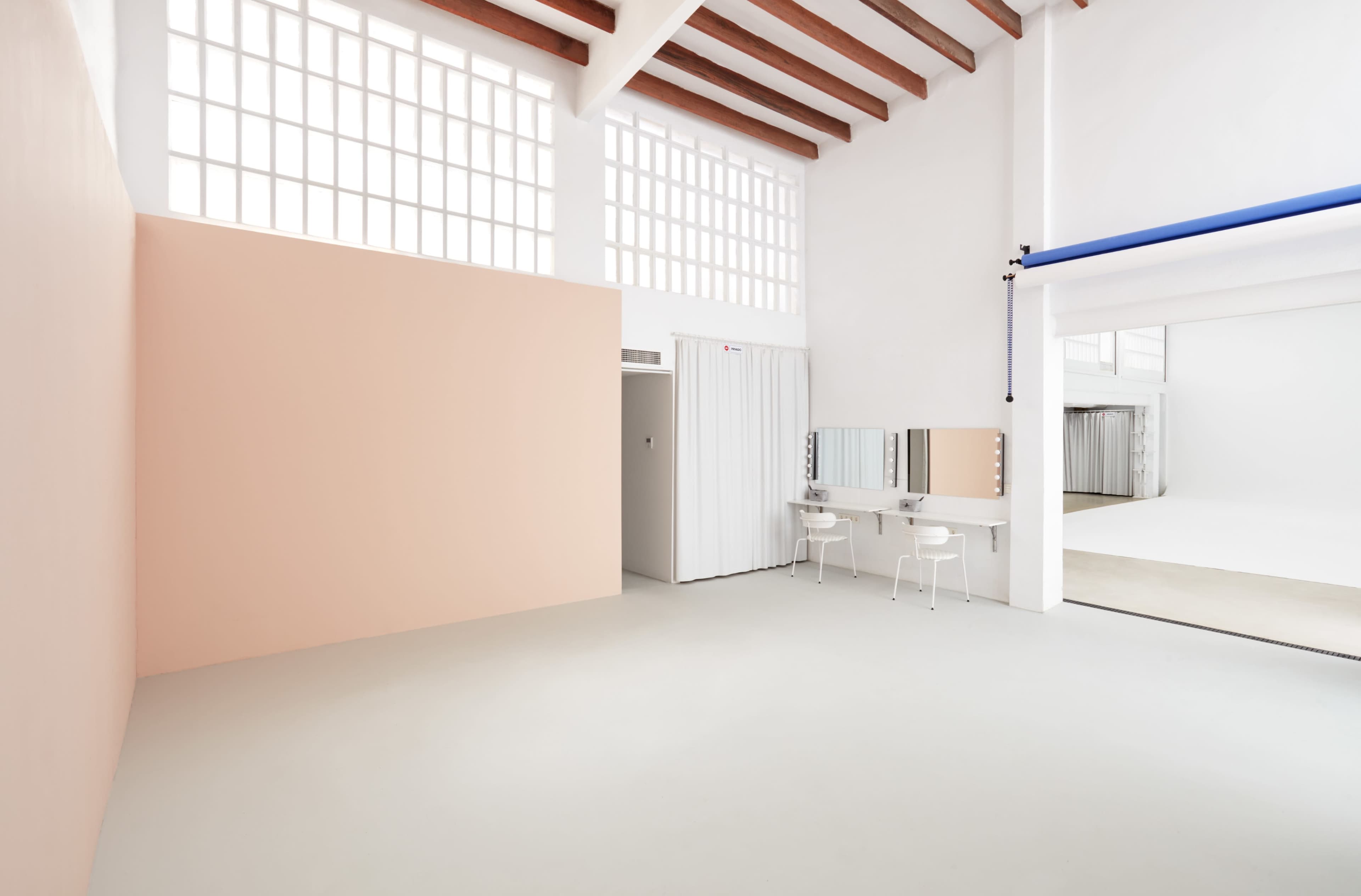 The image shows a spacious, well-lit studio with high ceilings, featuring a pale pink wall, a white dressing area with mirrors, and an adjoining empty space.