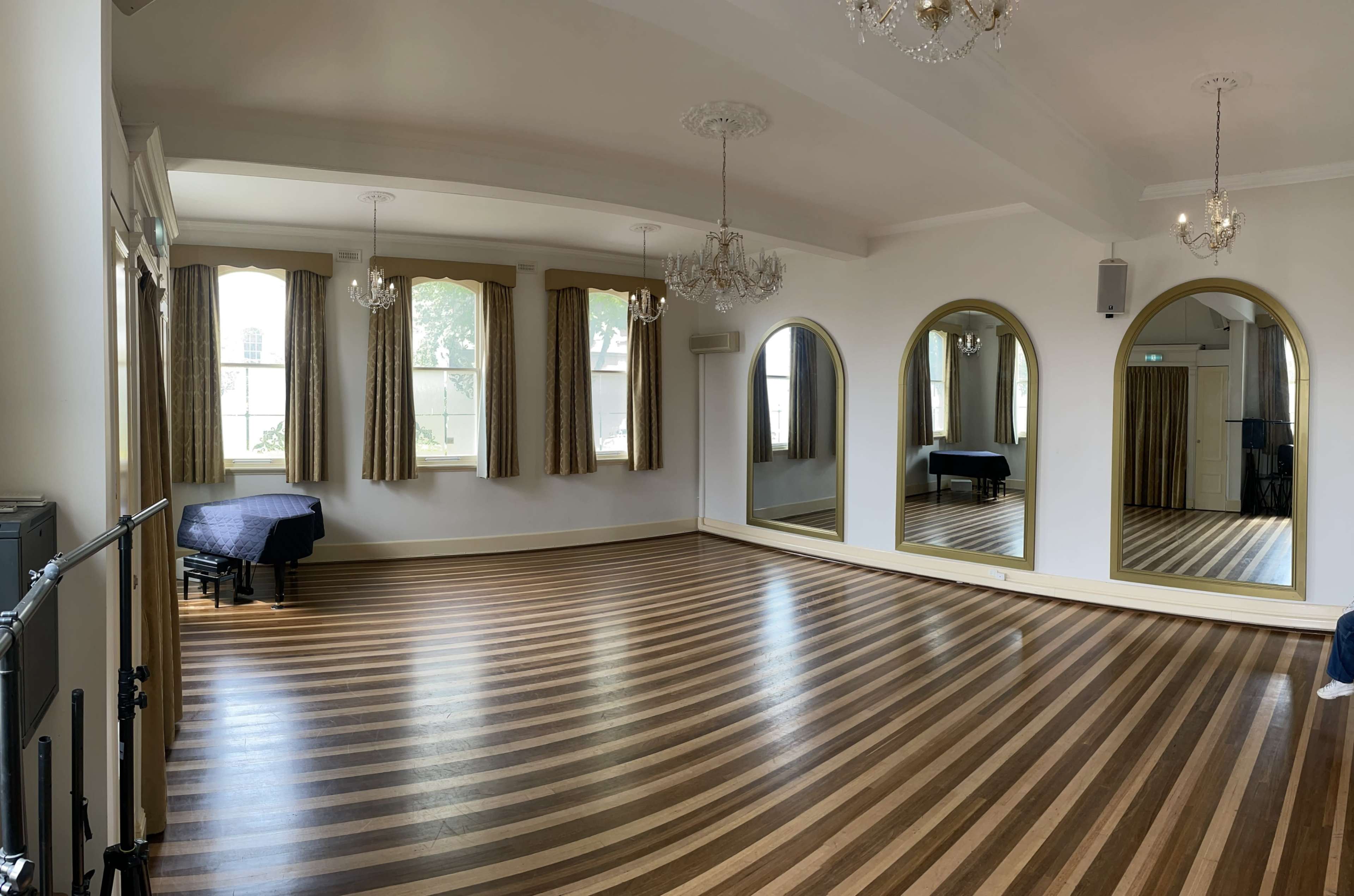 An empty, well-lit room with wooden floors, large mirrors, and two chandeliers hanging from the ceiling.