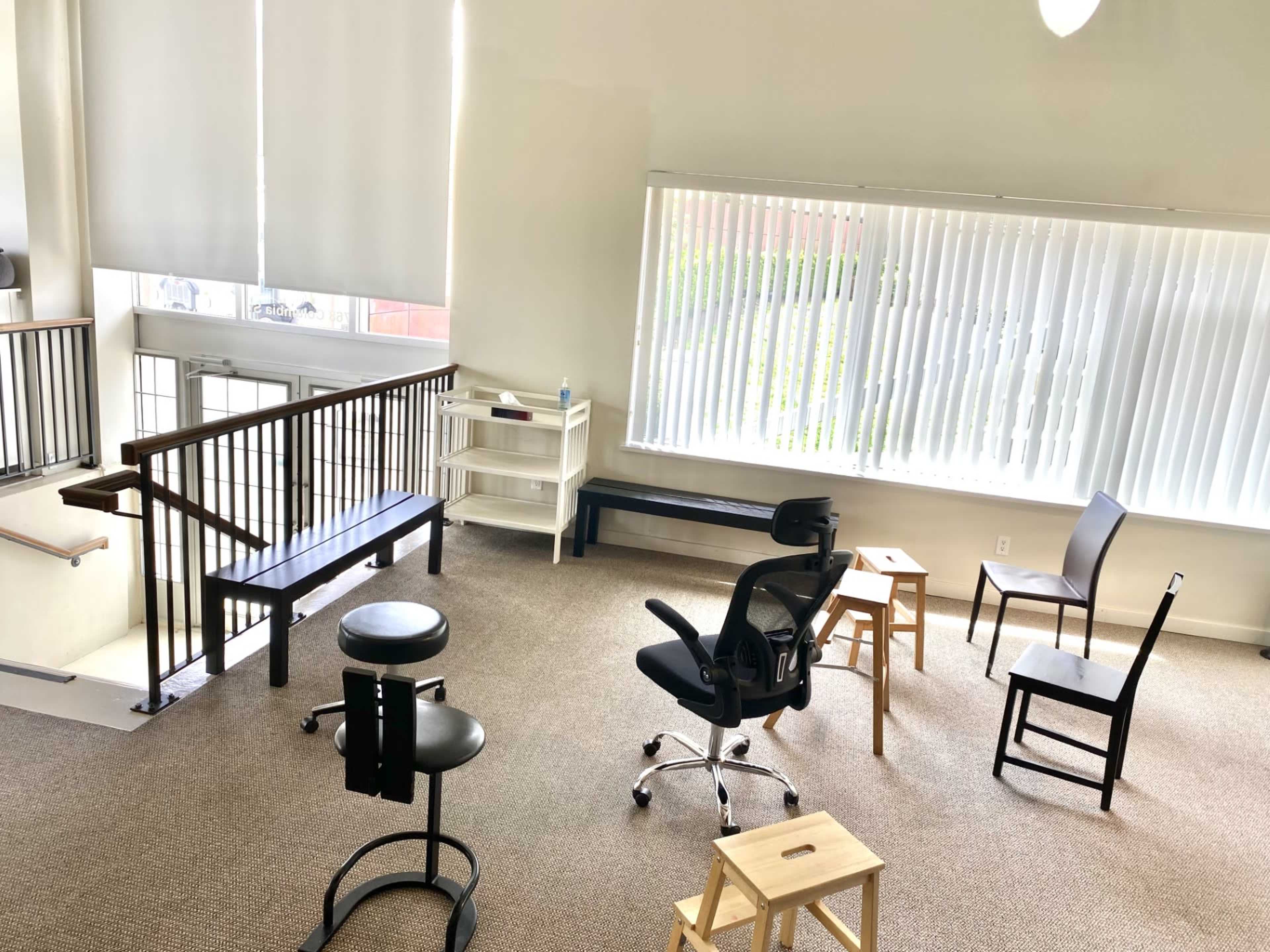 The image shows an empty room with a carpeted floor, several chairs, a small table, and large windows covered with blinds.