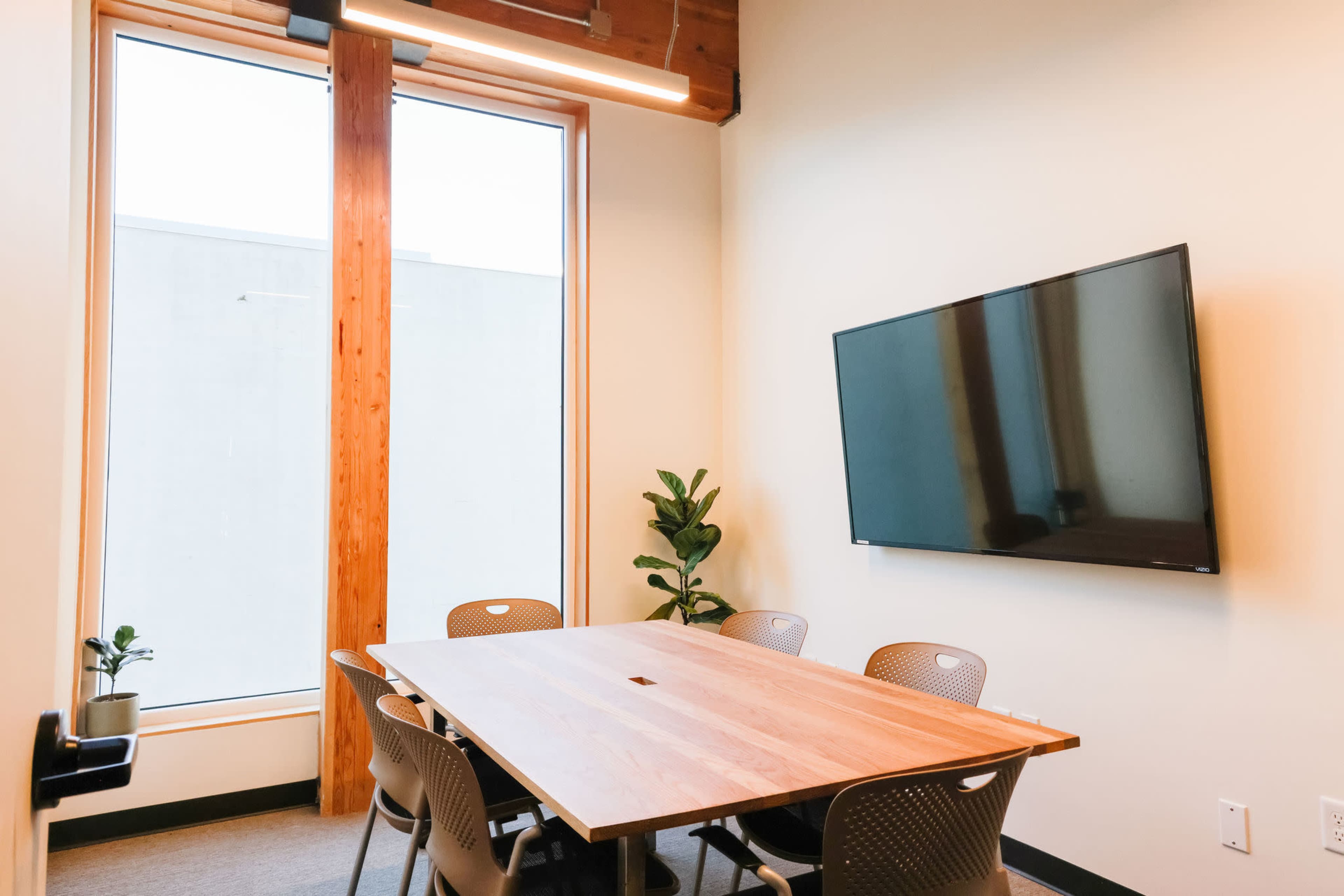 A well-lit meeting room features a large wooden table surrounded by six chairs, with a television mounted on the wall and a plant near the window.