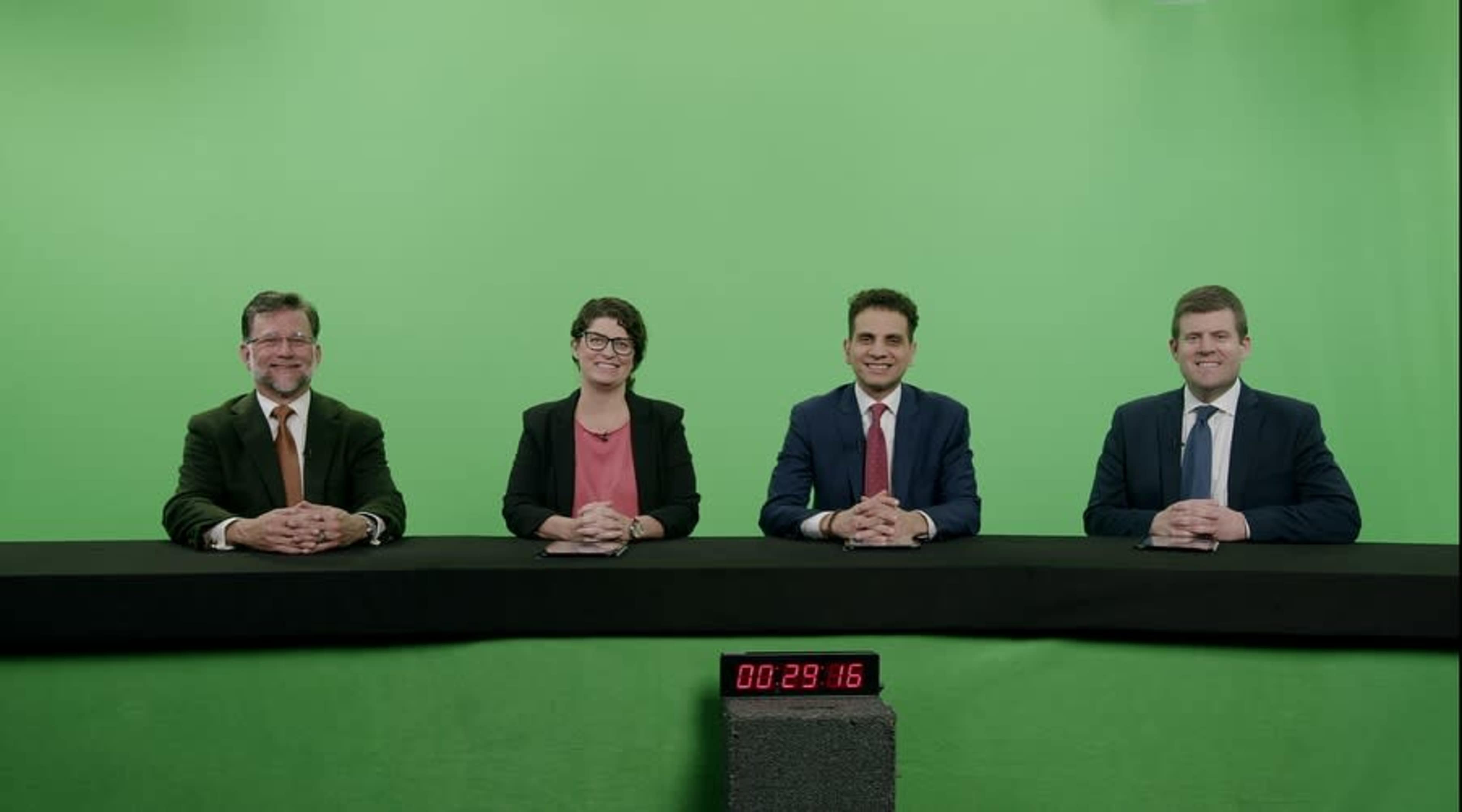 Four individuals sit at a table in front of a green screen, with a digital timer visible in the foreground.