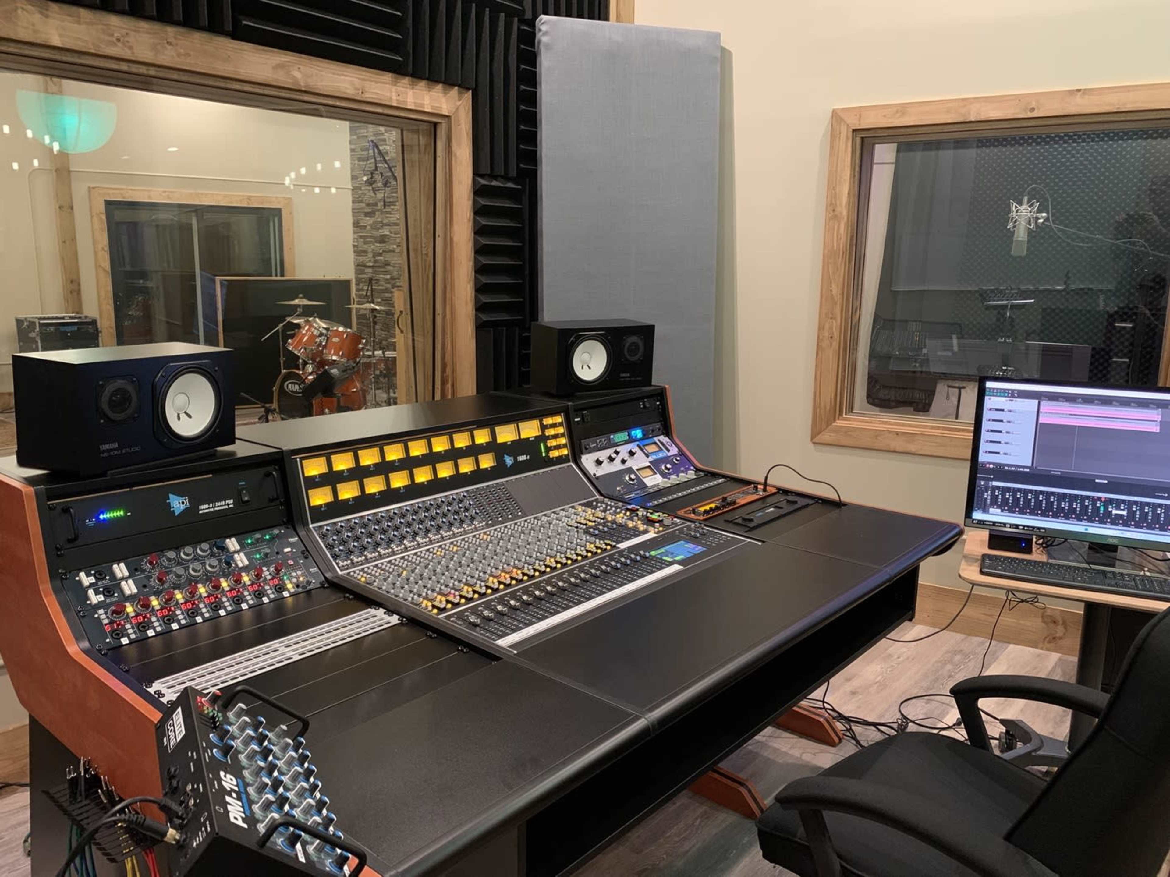 The image shows a music recording studio with a large mixing console, computer, and soundproof booths in the background.