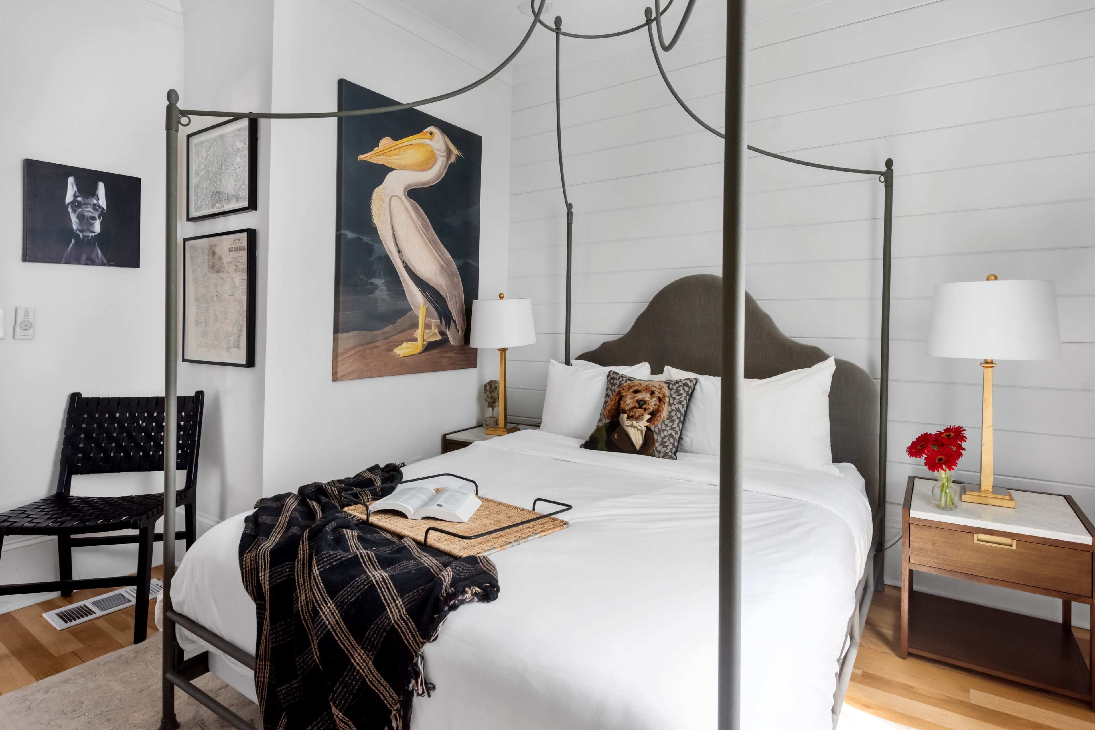 A canopied bed with white bedding is positioned in a bright room that features artwork on the walls and a small table with decorative items.