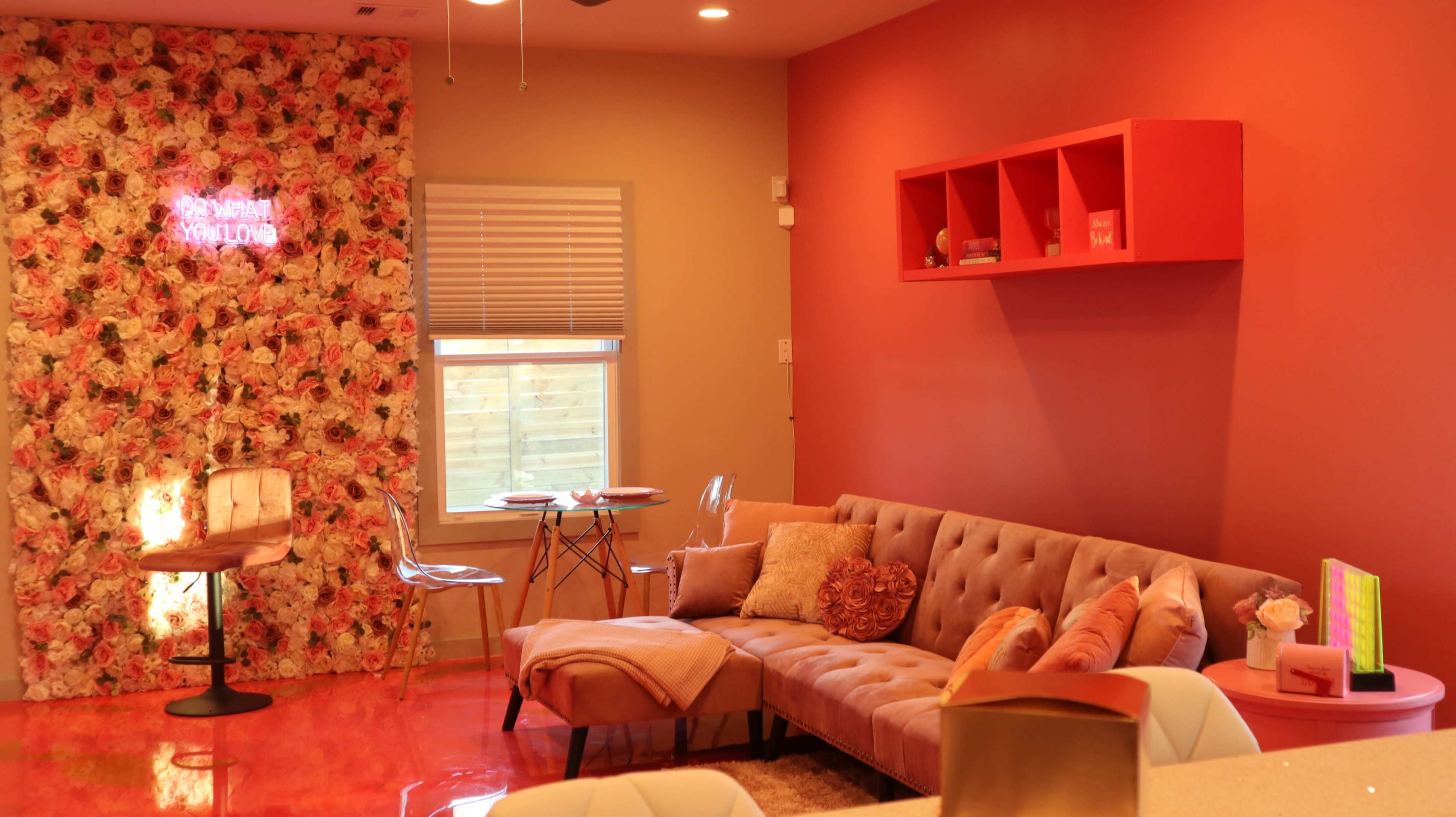 A cozy living room with a tufted sofa, a floral accent wall, and a small dining table set against a vibrant pink backdrop.