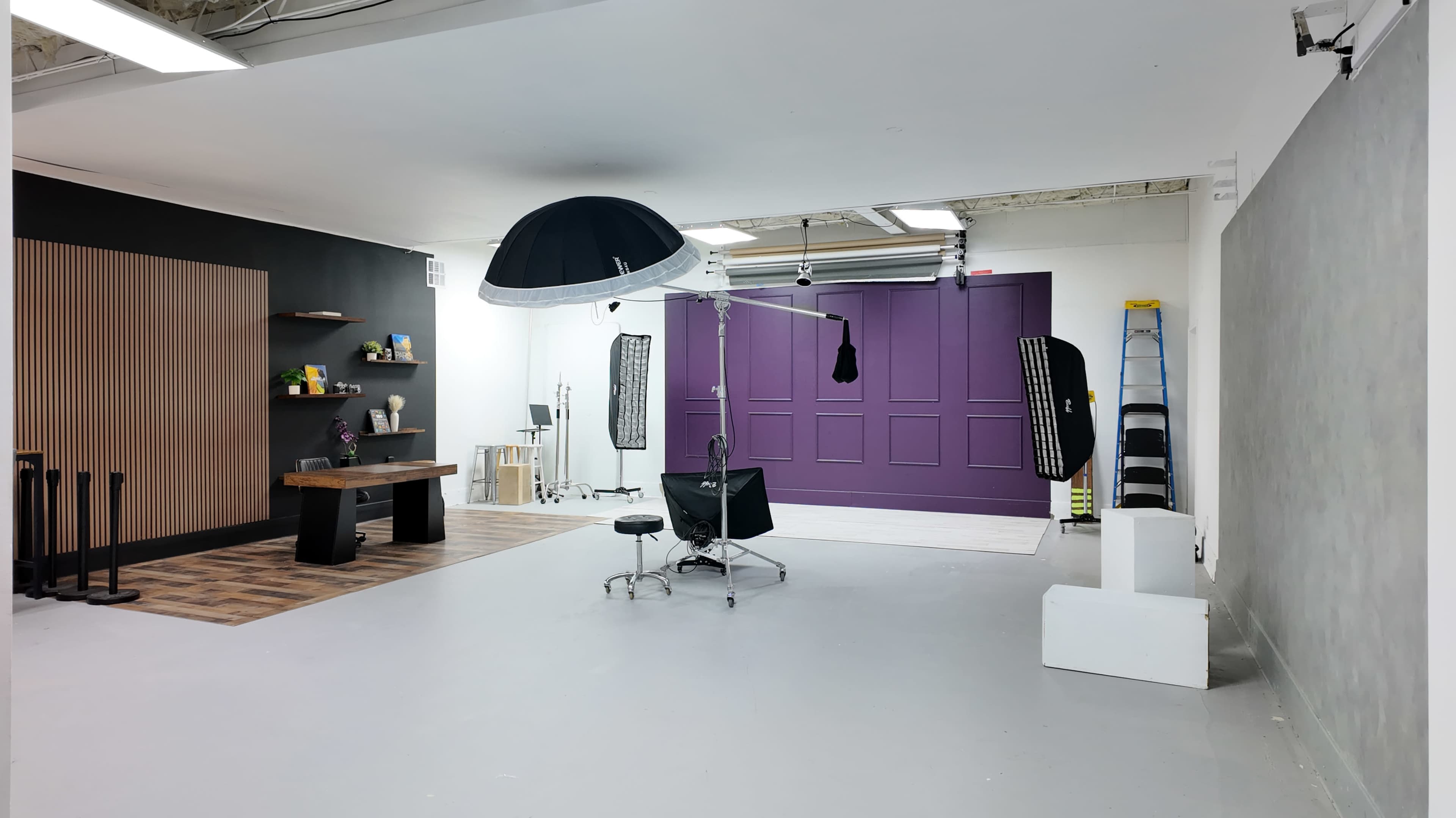 The image shows a photography studio featuring a large backdrop, a lighting setup, a work table with various items, and a purple wall.