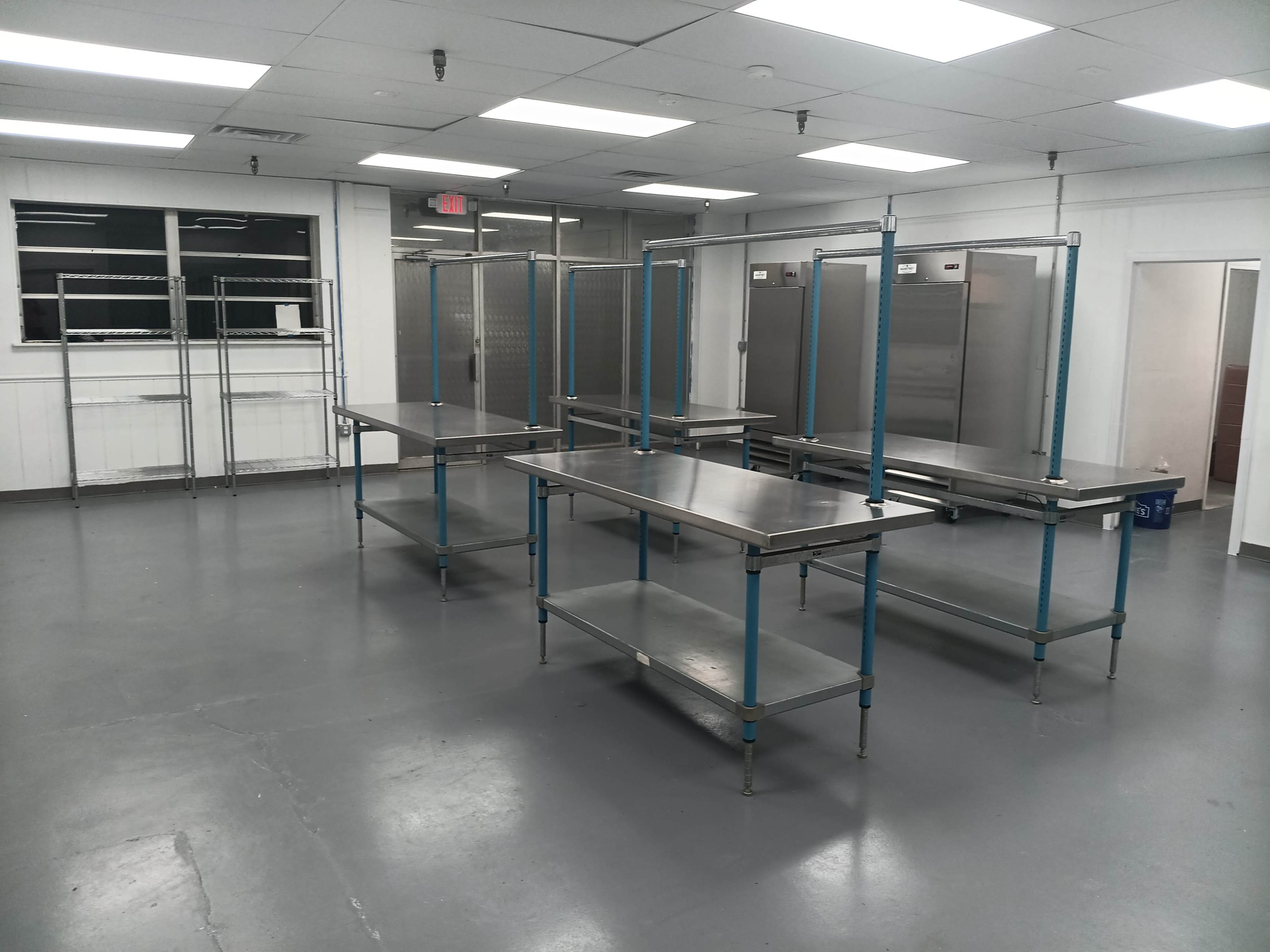 The image shows a spacious, empty kitchen area equipped with several metal tables and shelving units.