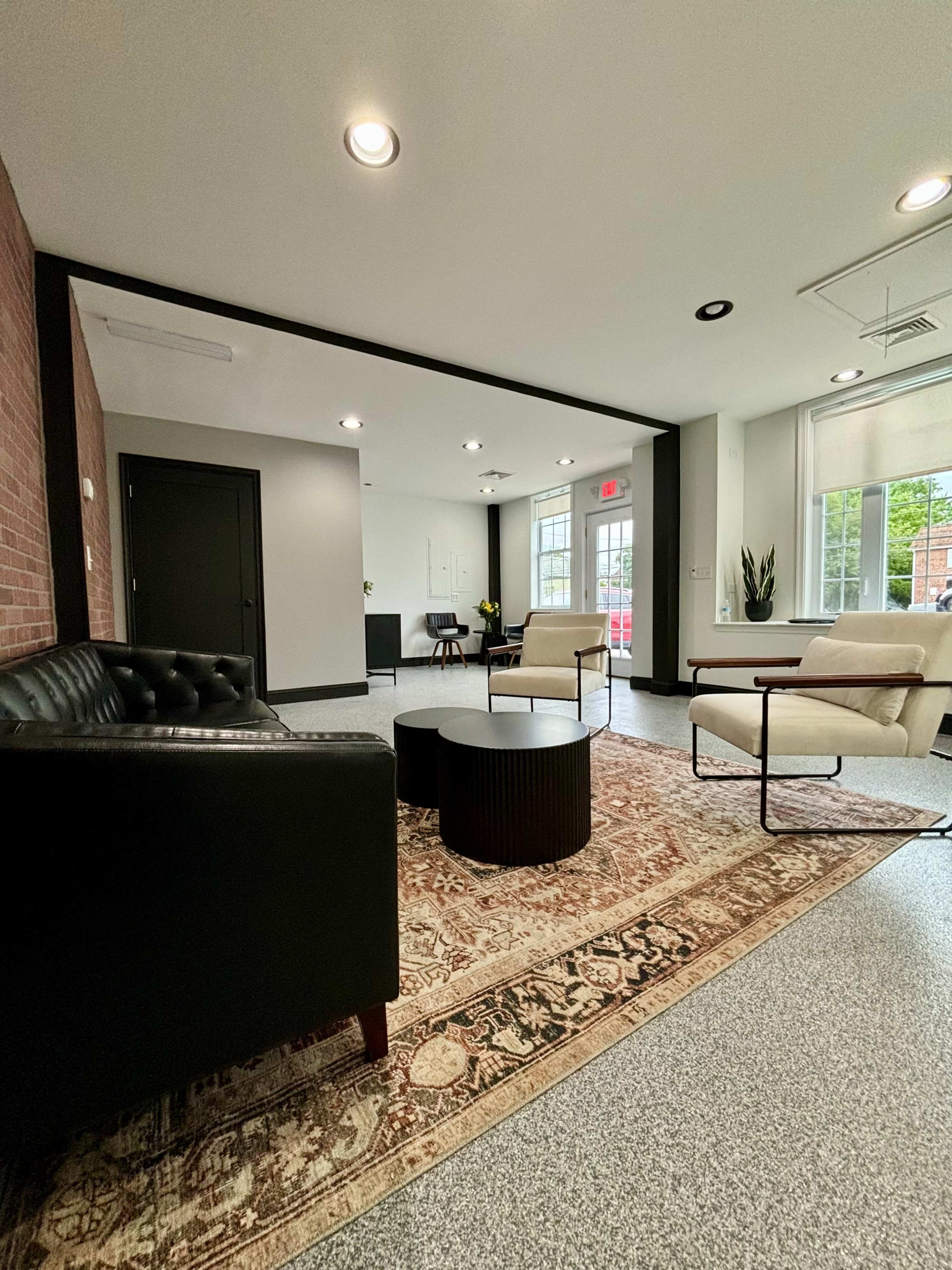 The image shows a modern waiting area featuring a black leather sofa, white armchairs, a round coffee table, and a patterned area rug, with large windows letting in natural light.