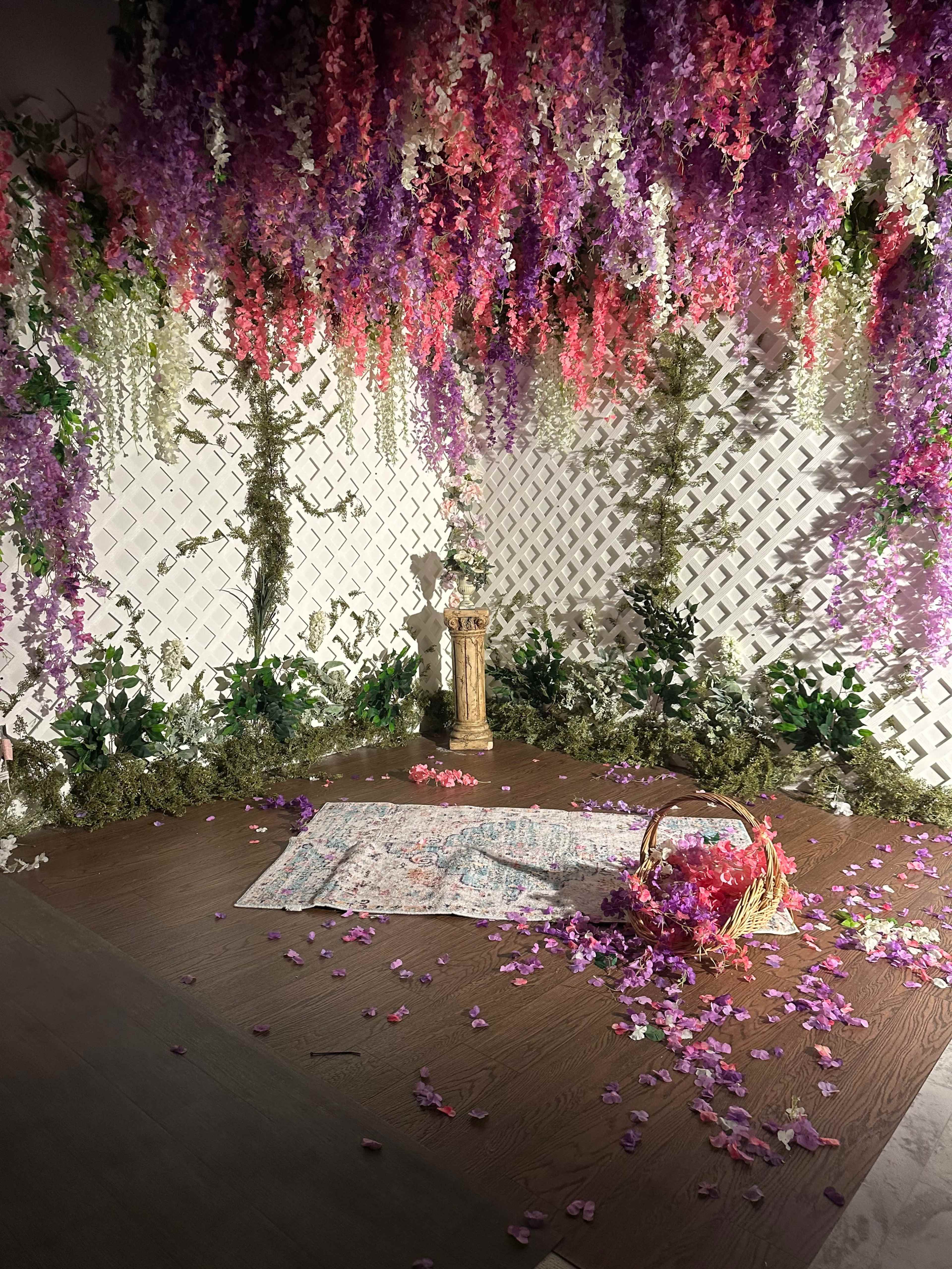 The scene features a space decorated with hanging purple and pink flowers, a wooden pedestal in the center, and a patterned blanket on the floor surrounded by scattered petals and a woven basket.