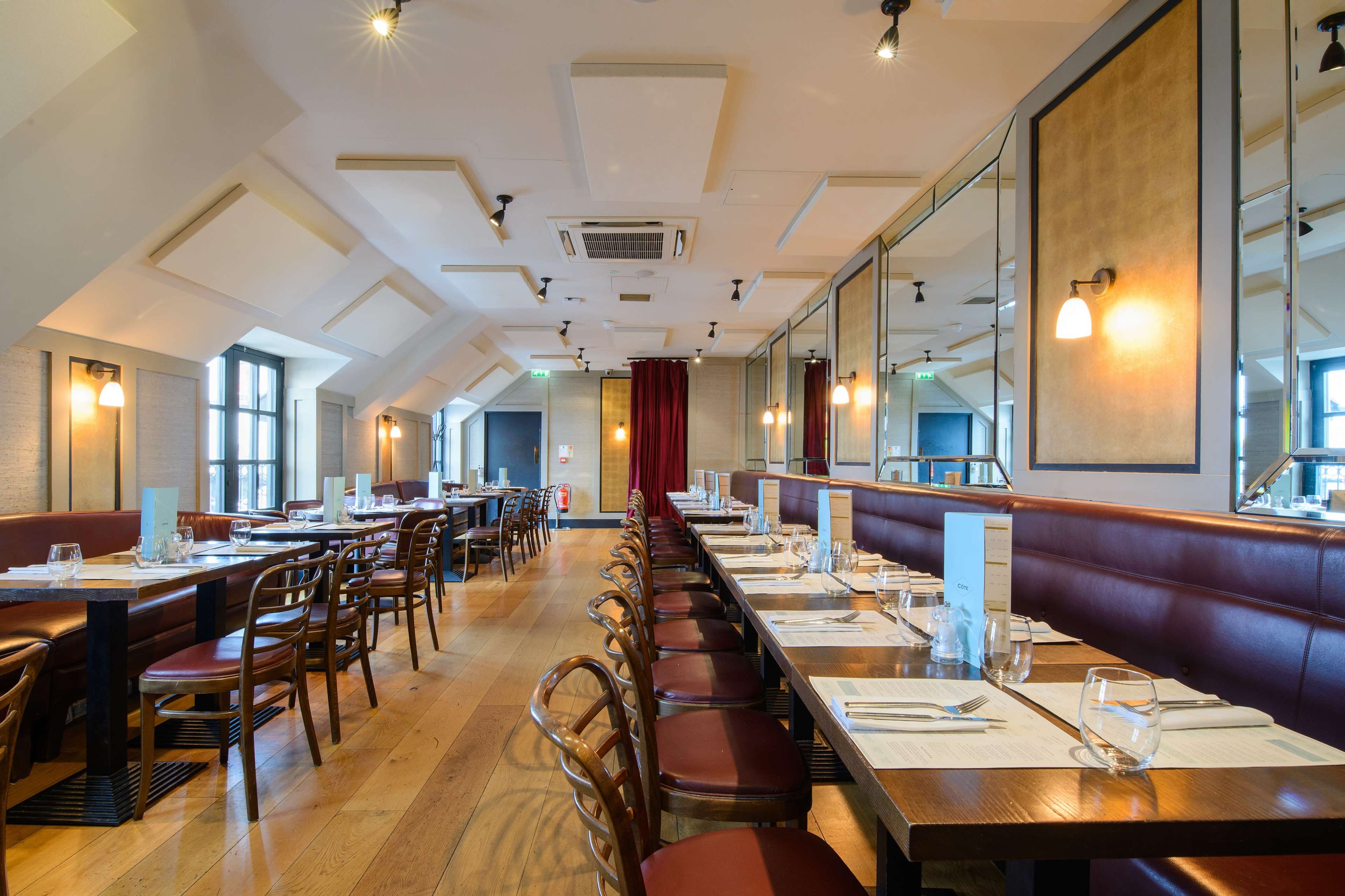 The image shows a modern restaurant interior with wooden floors, rows of tables lined with chairs, and large windows allowing natural light.