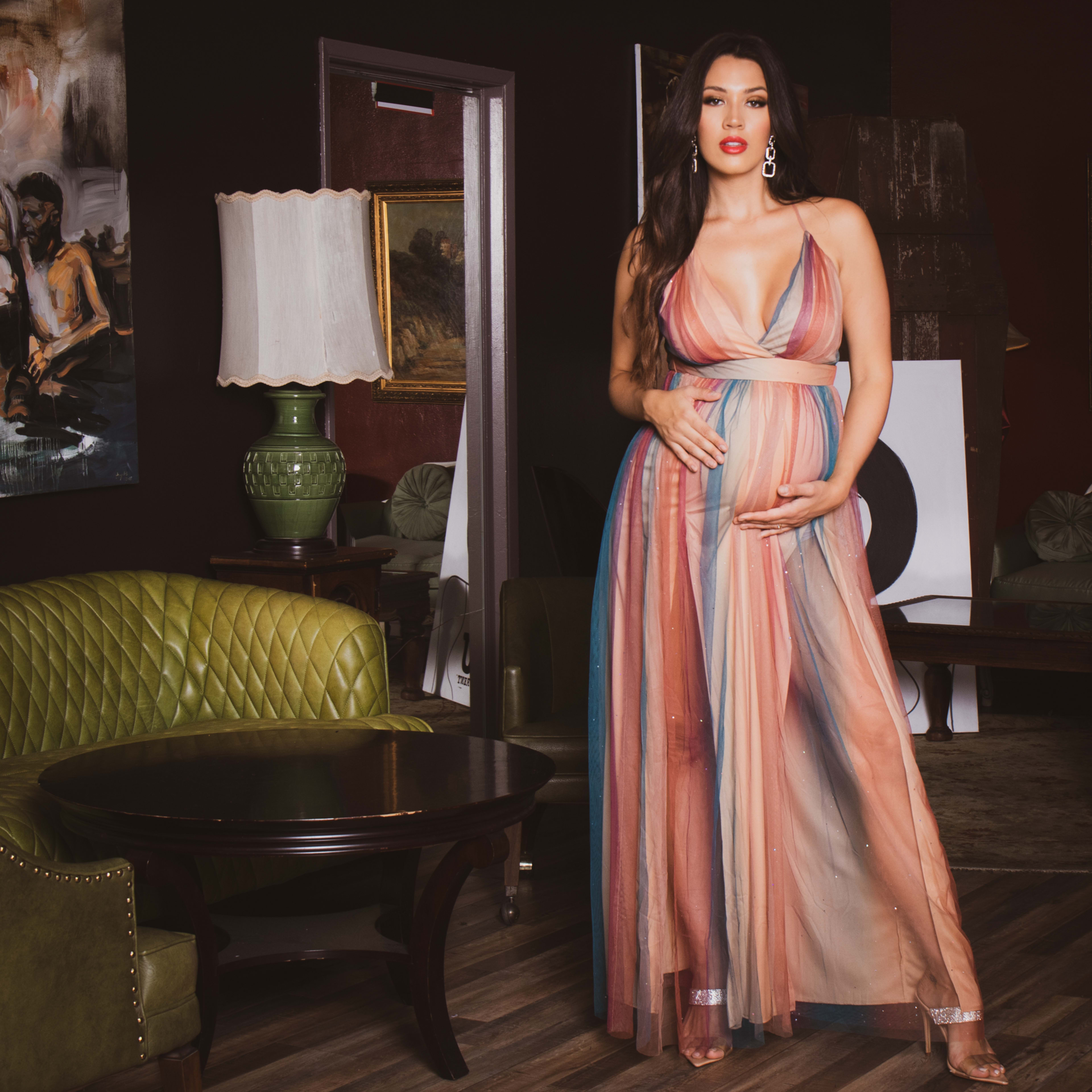 A maternity photoshoot of a woman in a dress holding her belly in a living room.