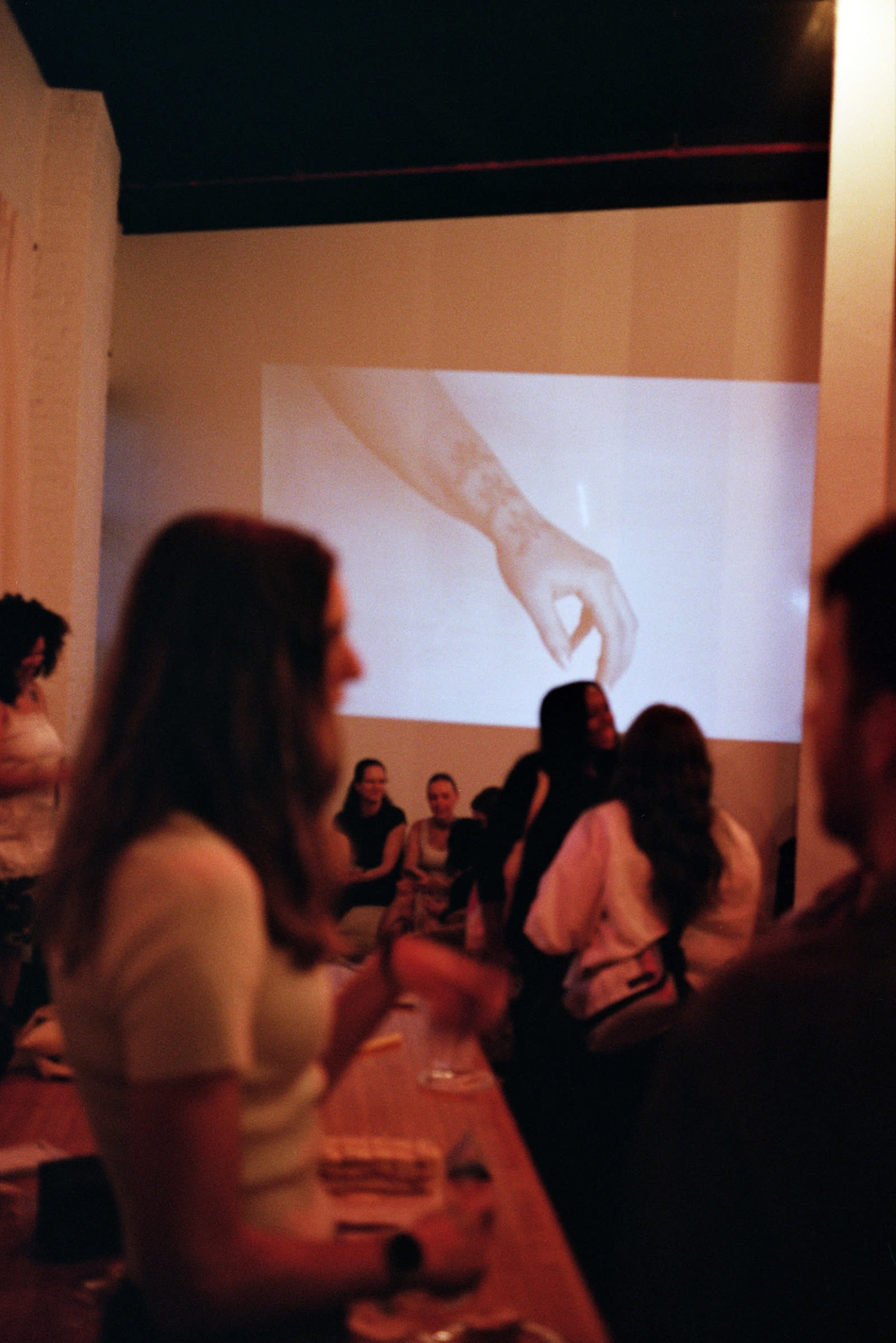 The image shows a dimly lit gathering in an art space, with people socializing in the foreground and a video projection of a hand with tattoos in the background.