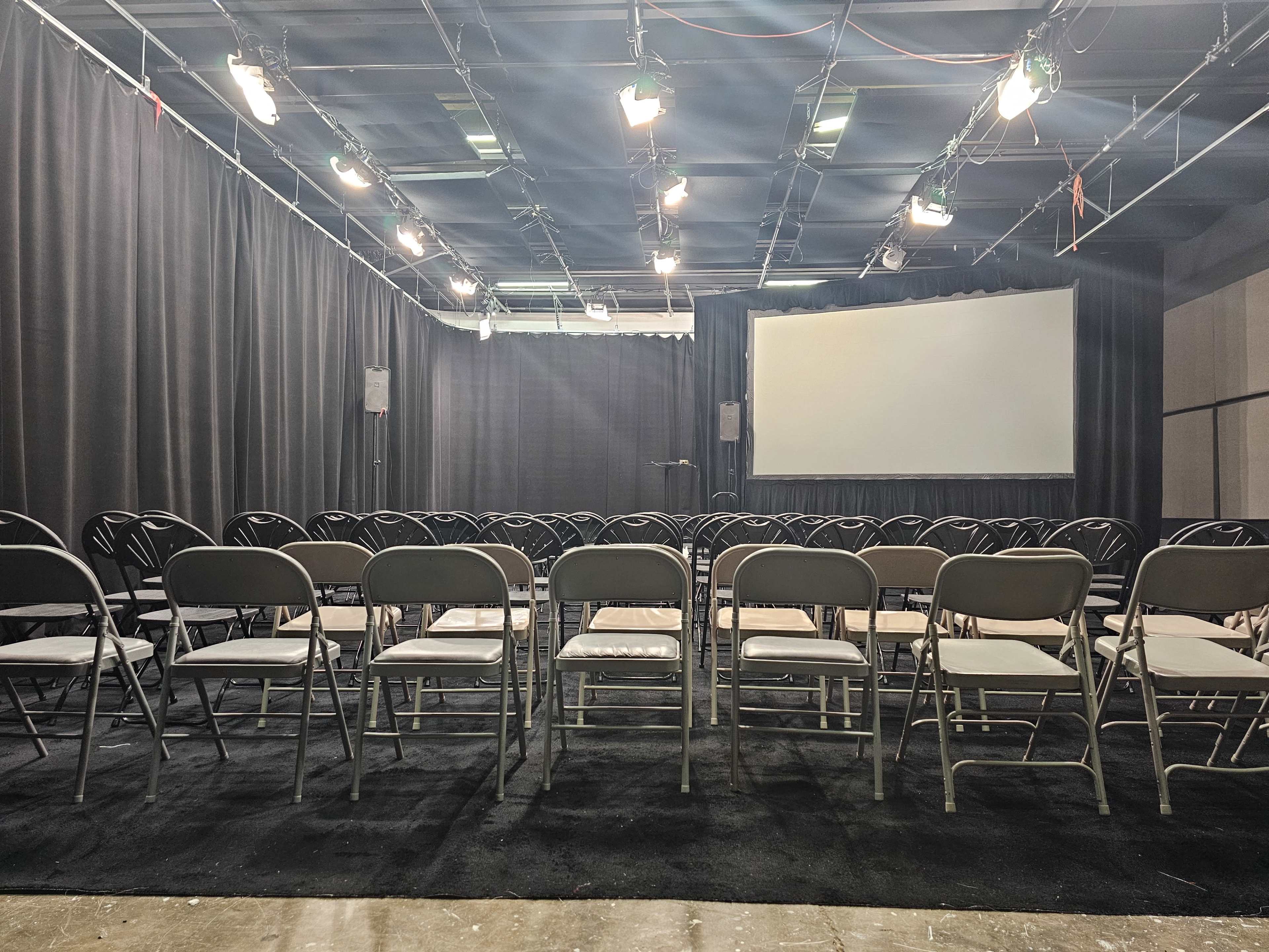 The image shows a dimly lit auditorium set up with rows of folding chairs facing a blank screen at the front.