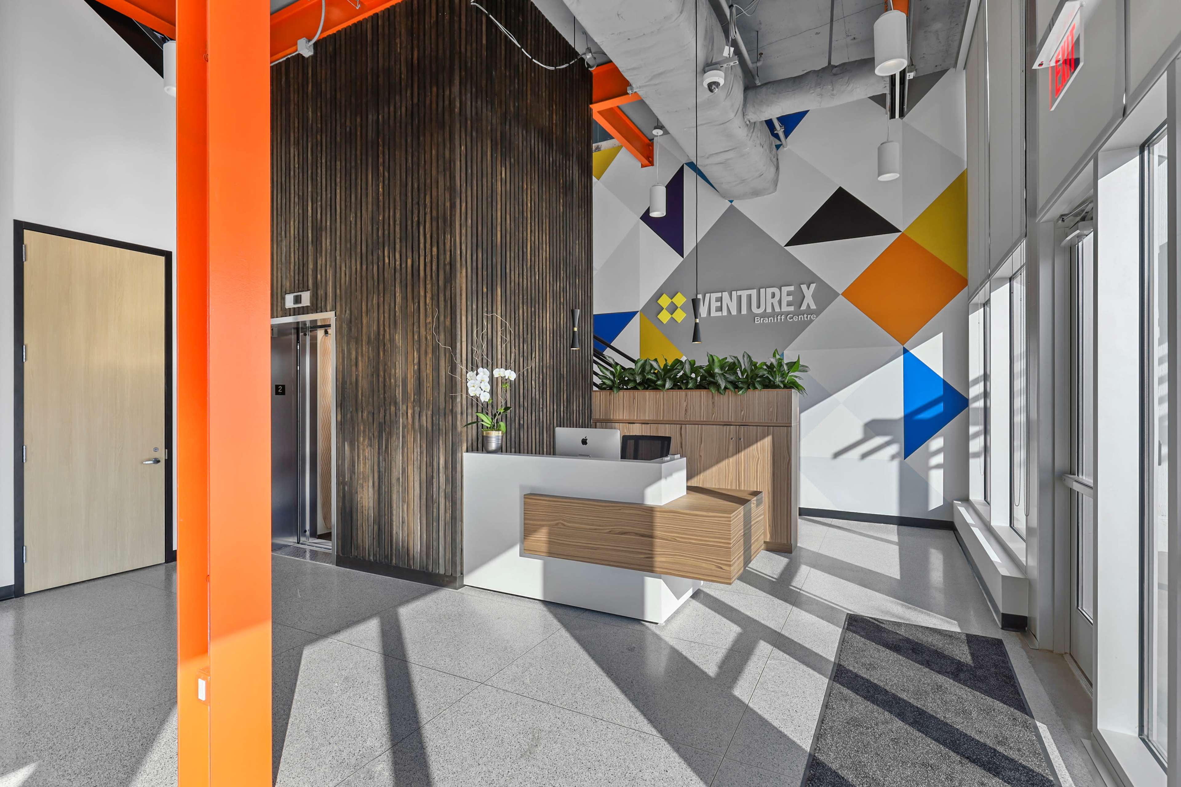 The image shows a modern reception area with a wooden front desk, an elevator, and a colorful wall featuring the logo of Venture X.