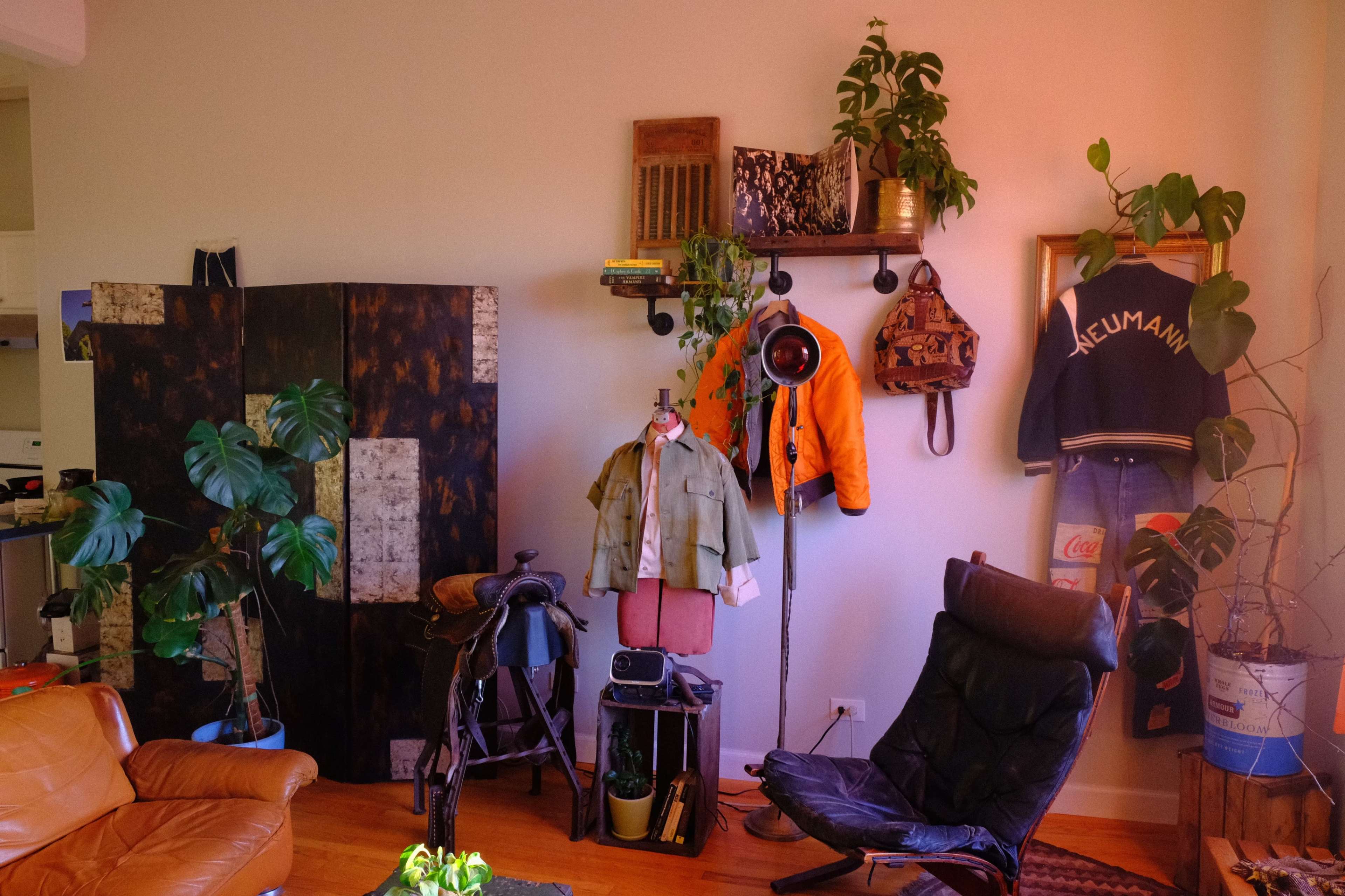 The image shows a cozy living space featuring a mix of vintage clothing, plants, and rustic decor against a light-colored wall.