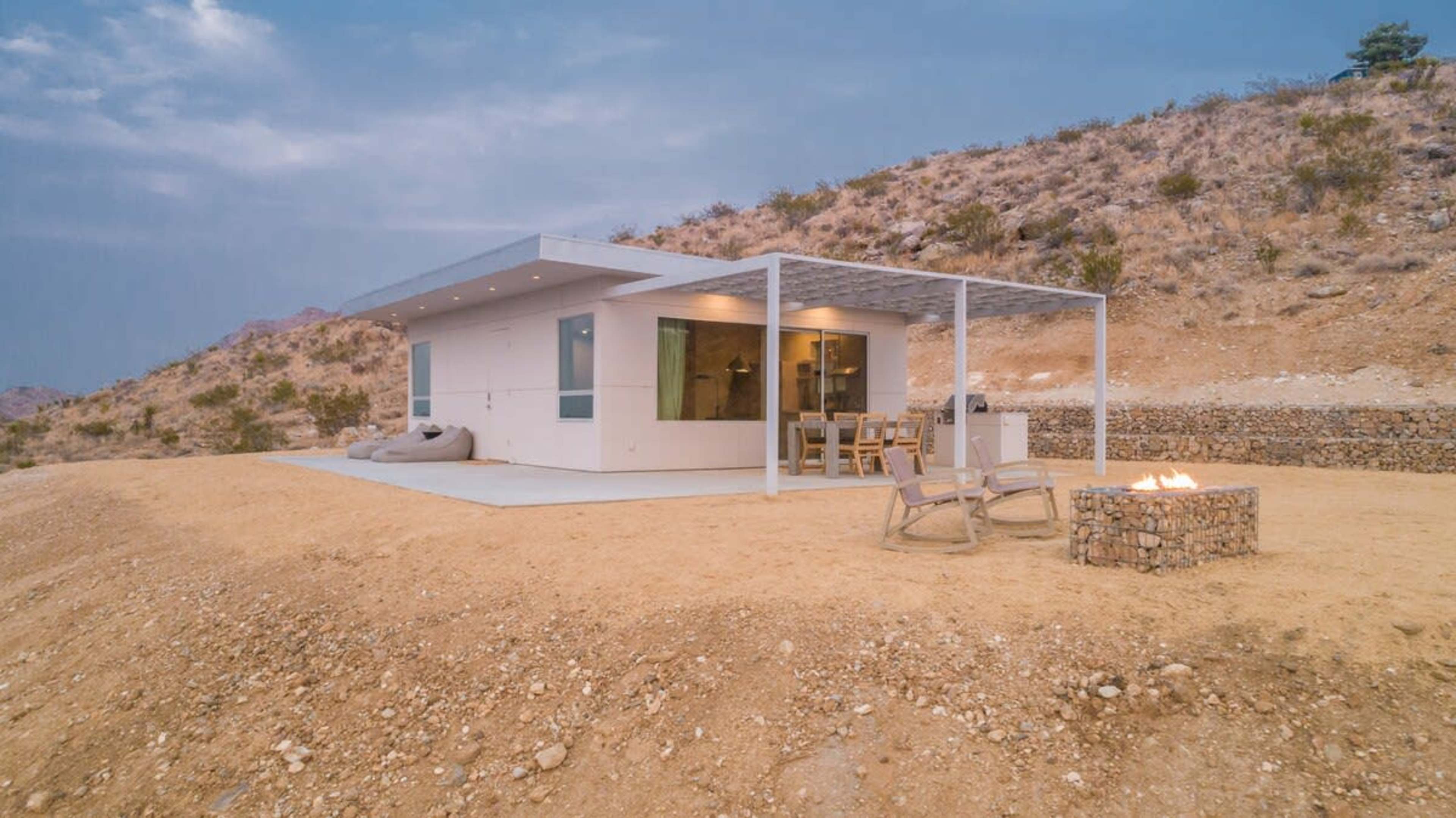 A modern house with a patio and outdoor seating is situated on a hillside, surrounded by dry, rocky terrain.