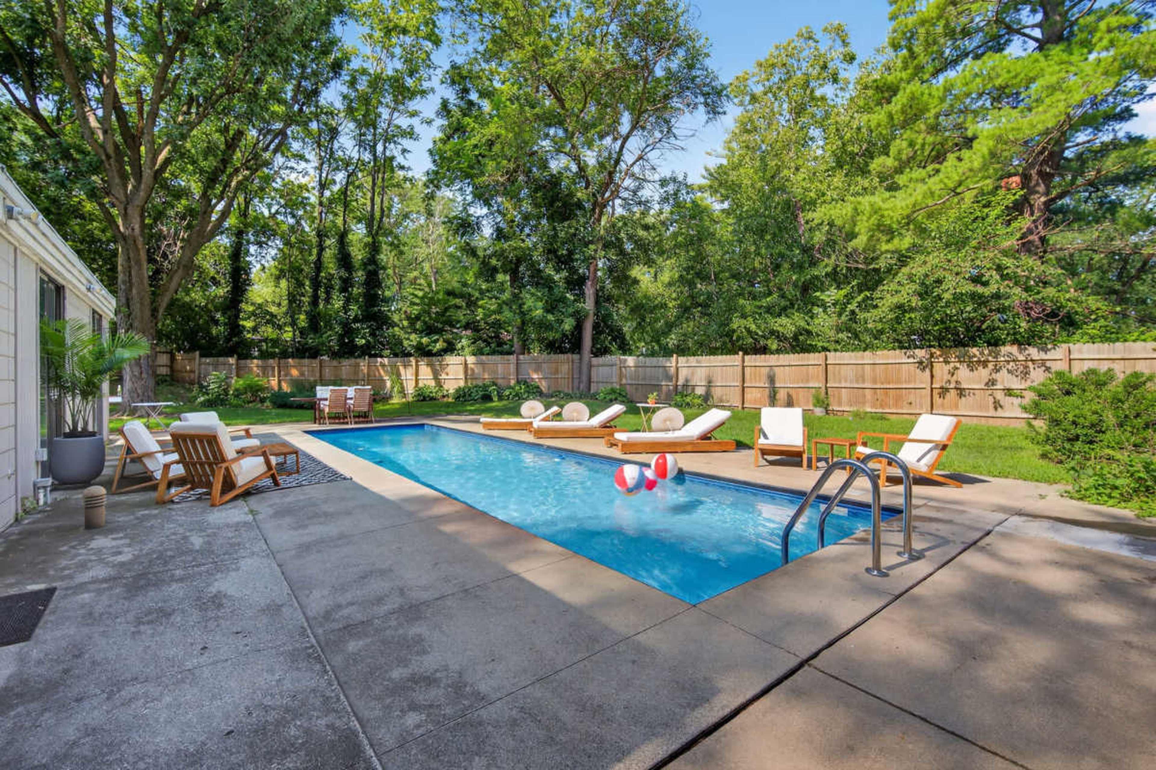 The image shows a backyard pool surrounded by lounge chairs and a wooden deck, with a fence and trees in the background.