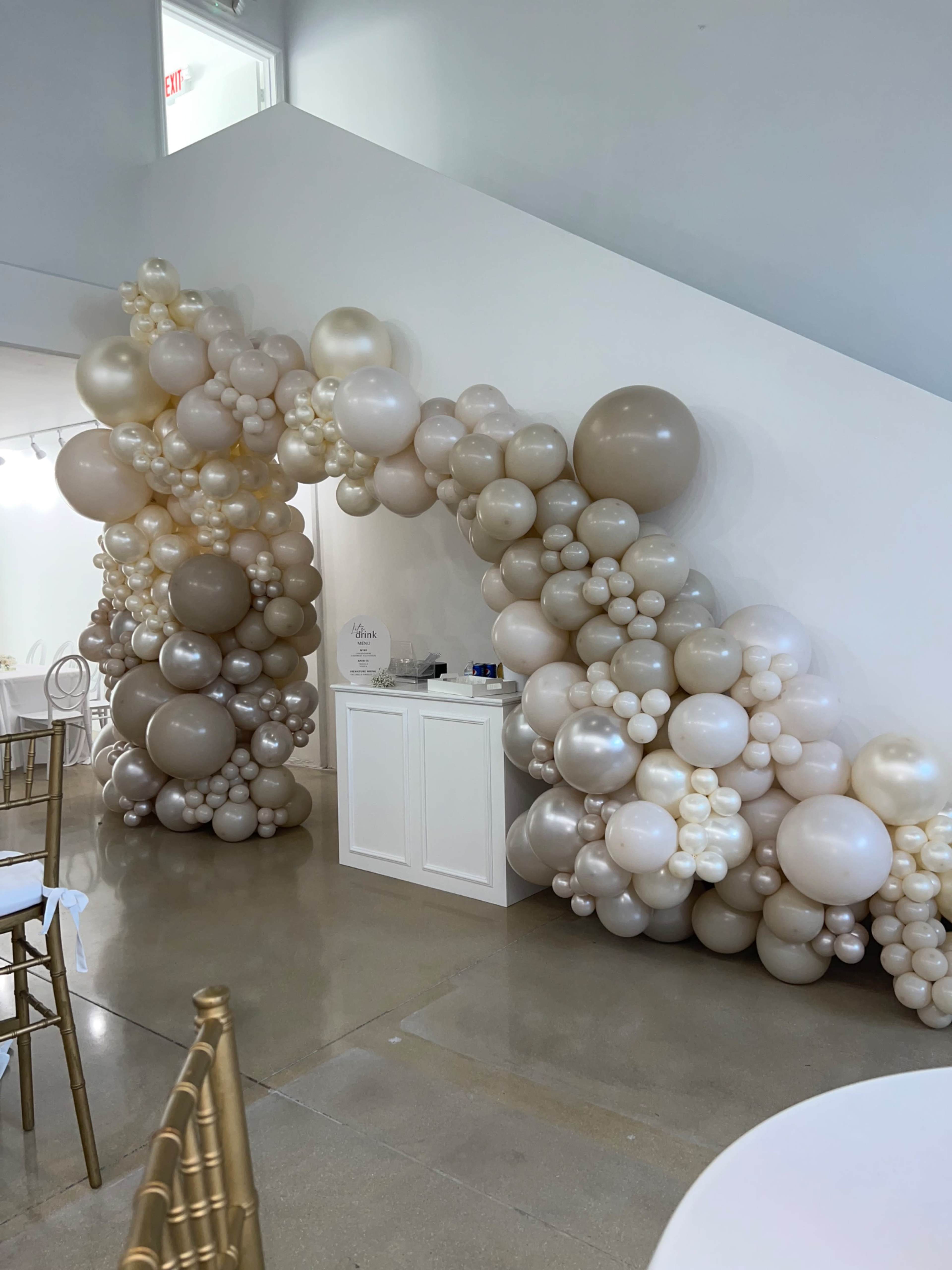 A decorative arch made of various sizes of beige and cream balloons is positioned near a white service table in a well-lit venue.