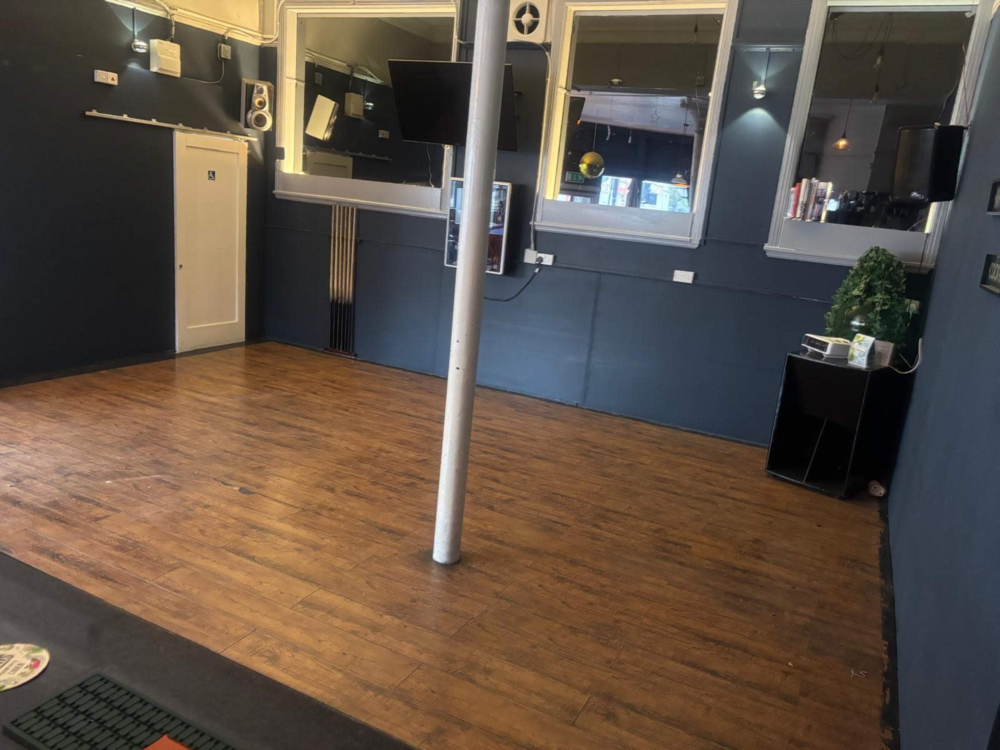 The image shows an empty room with wooden flooring, a single pole in the center, and large windows with mirrors on one wall.