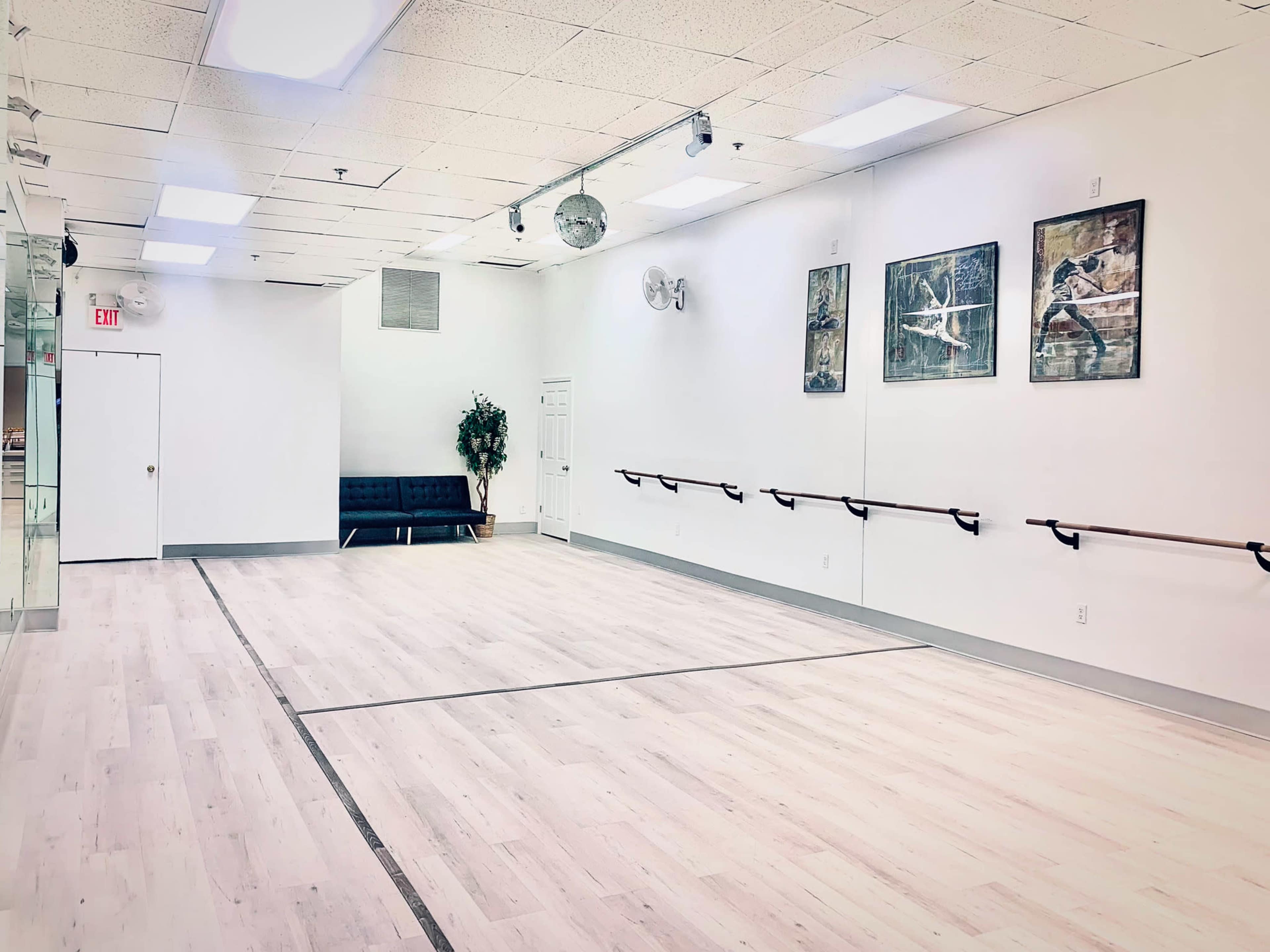 The image shows a spacious, empty dance studio with wooden flooring, mirrored walls, and a ballet barre along one side.