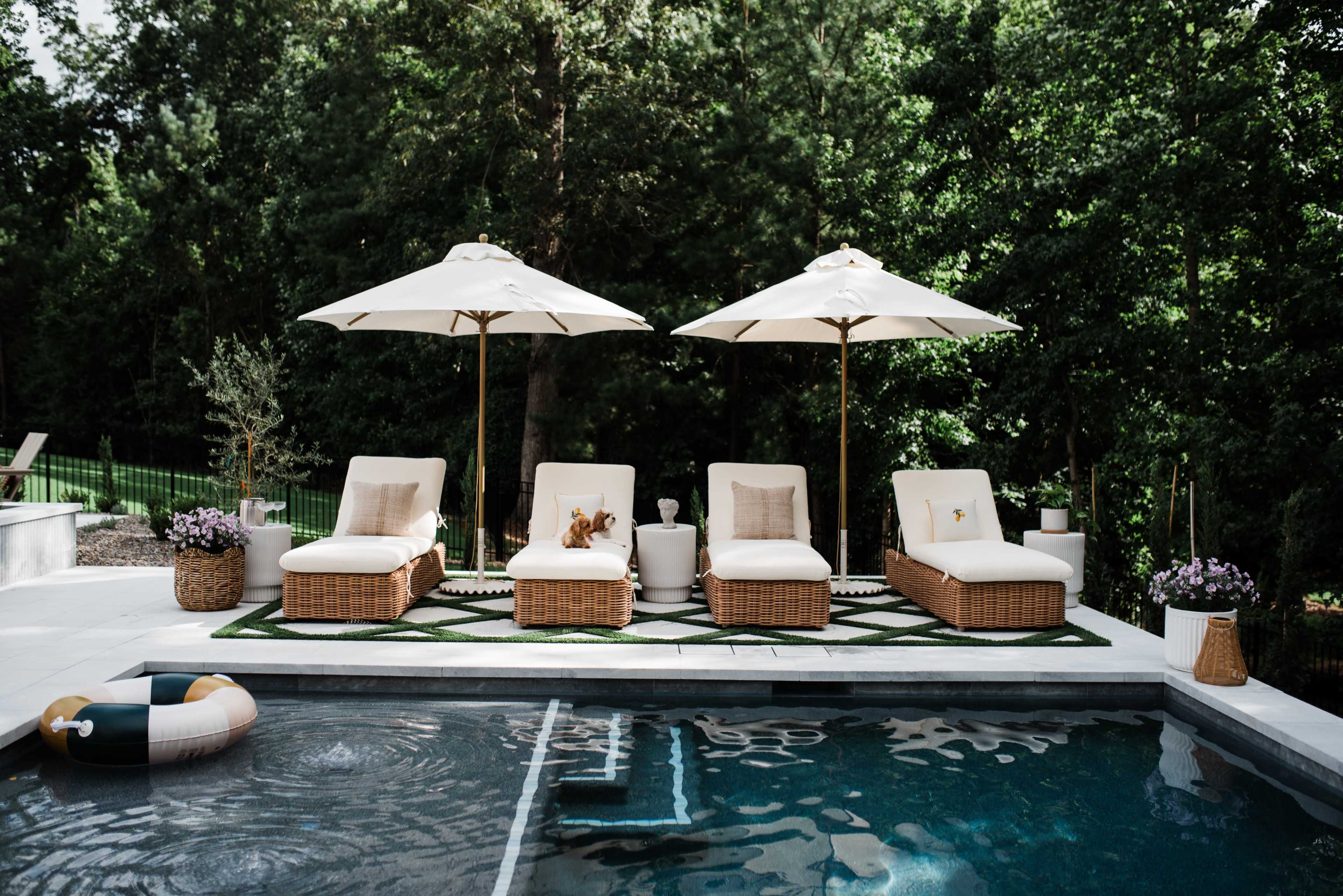 The scene features four lounge chairs with white cushions and umbrellas around a swimming pool, set against a backdrop of green trees and a manicured lawn.