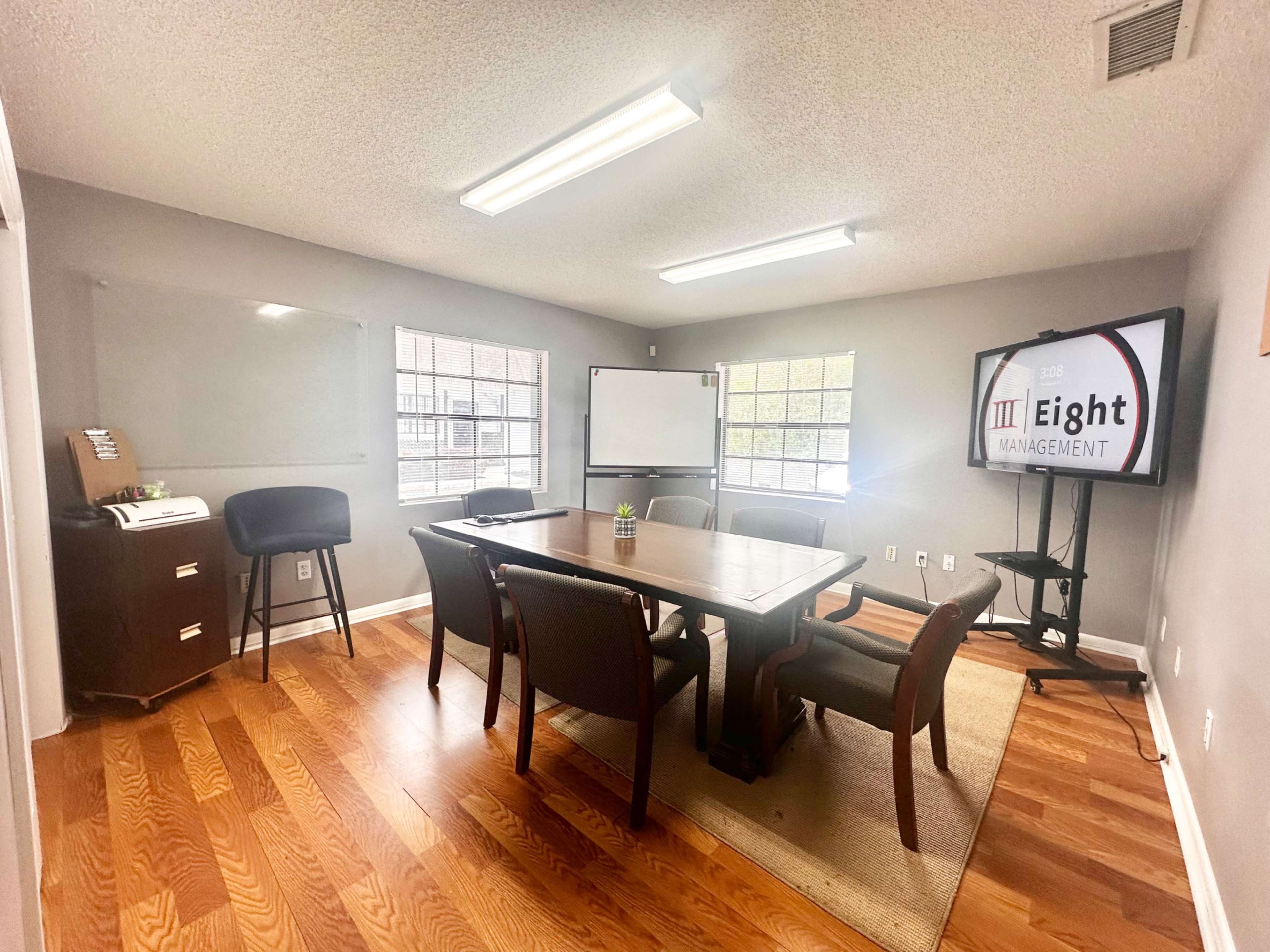 The image shows a meeting room with a large wooden table surrounded by several chairs, a whiteboard, a television on a stand, and two windows letting in natural light.