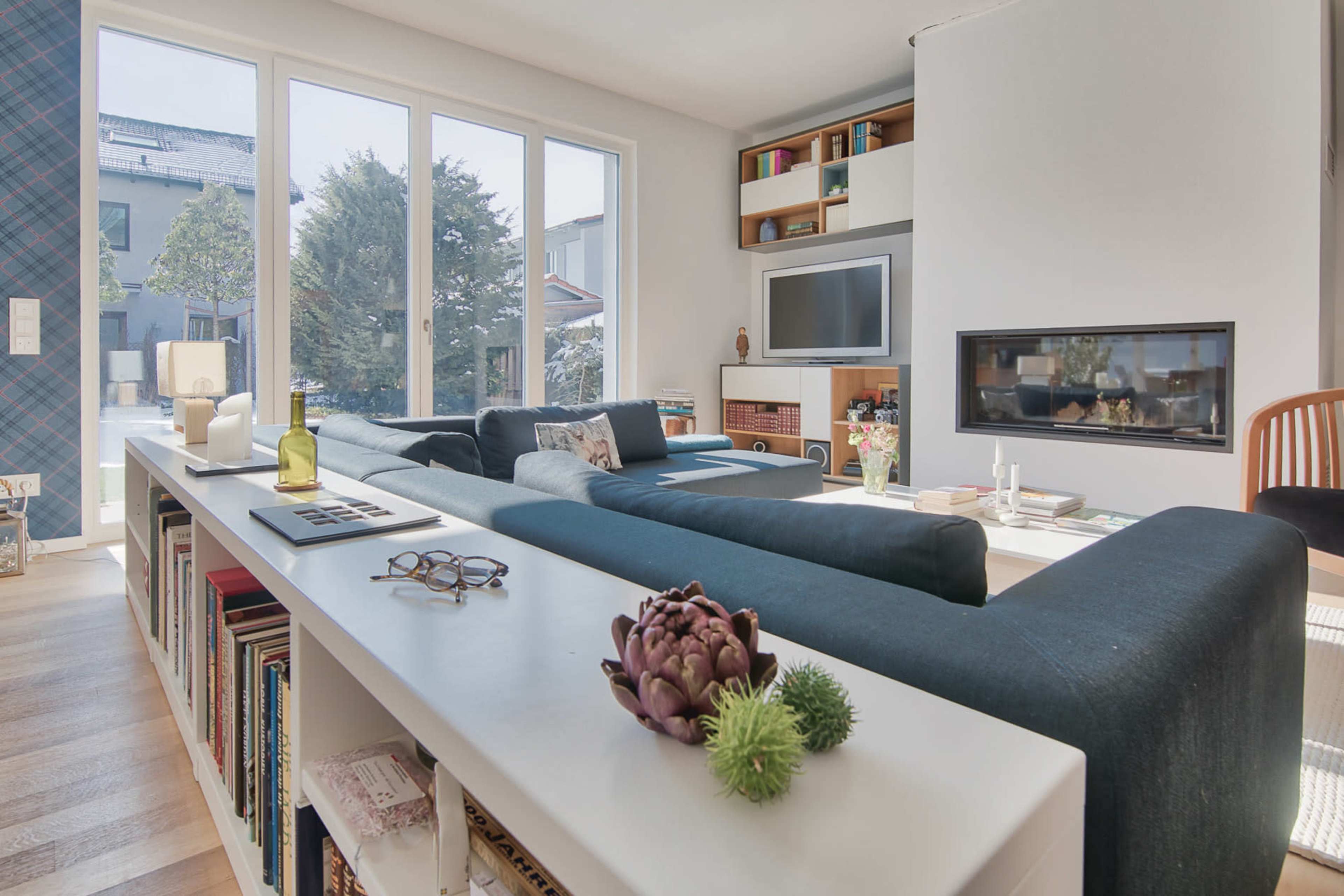 The image shows a modern living room featuring a sectional sofa, a television mounted on the wall, a fireplace, and large windows that provide natural light.