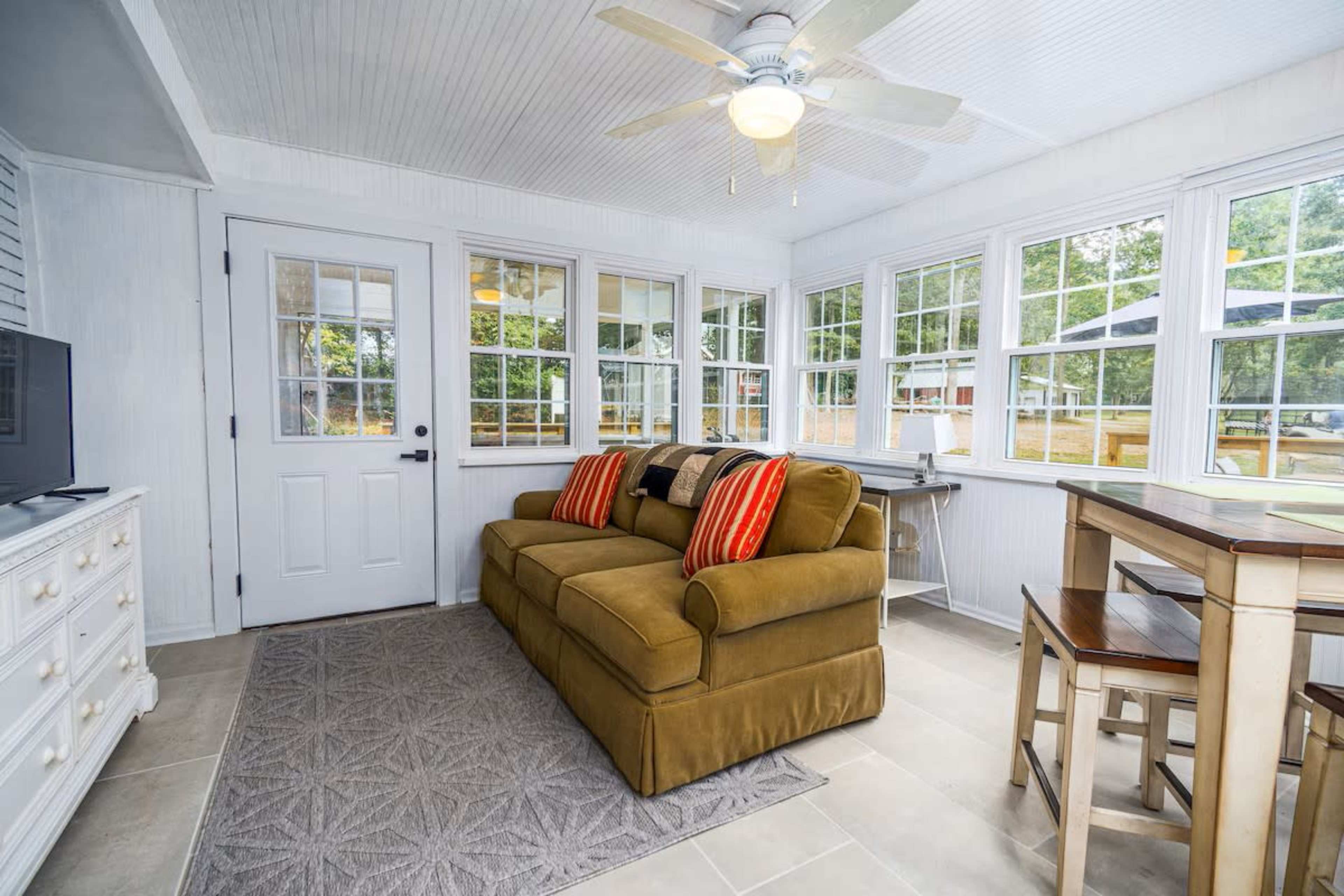 The image shows a sunroom featuring a brown couch with decorative pillows, a small dining table, and multiple windows allowing natural light to fill the space.