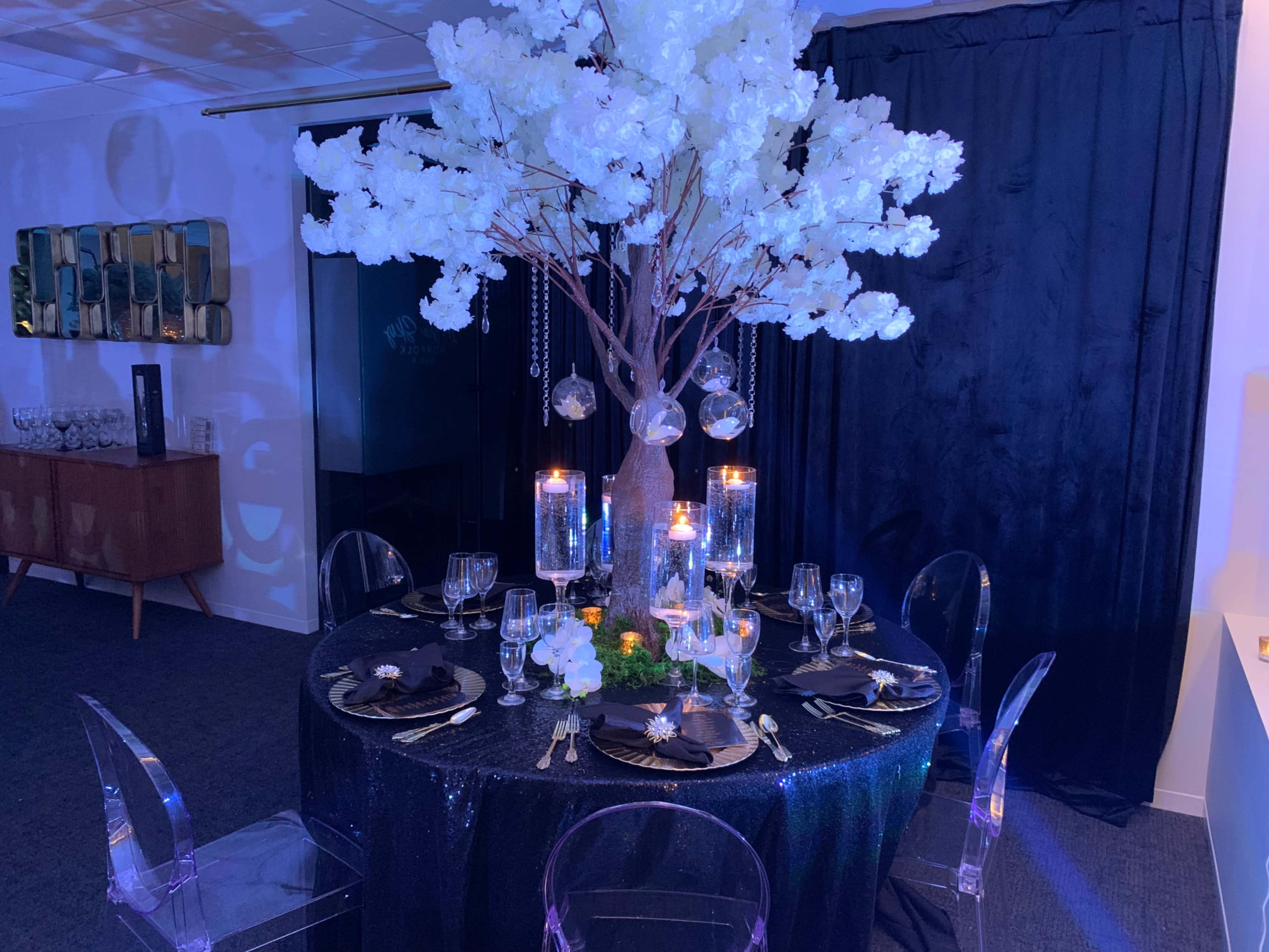 A round table is set with clear chairs, decorated with a tall white floral centerpiece and surrounded by candles, against a backdrop of dark drapery.